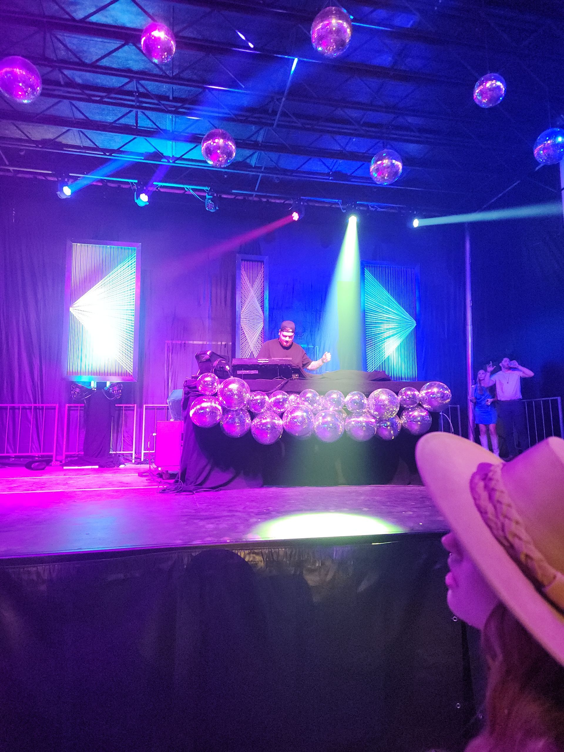 DJ on a neon-lit stage with pink and blue lights, disco balls, and a crowd in front