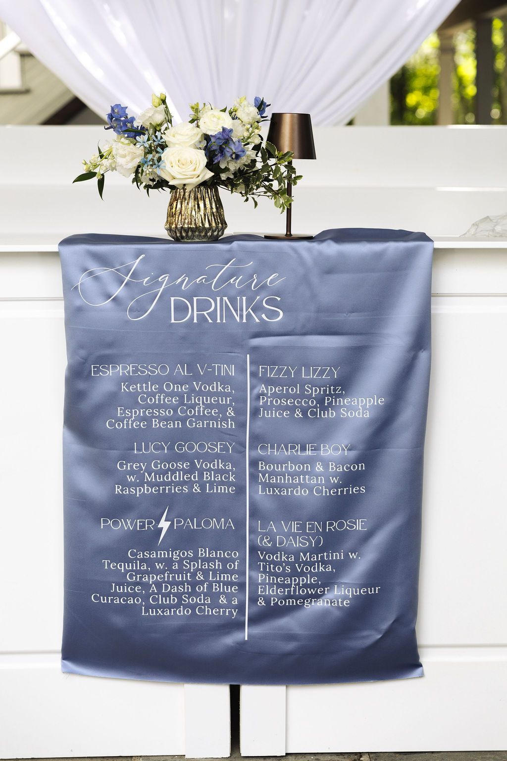 Blue wedding drink menu sign on a white mantel with flowers and curtain backdrop