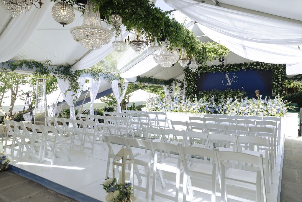 White outdoor wedding reception tent with rows of chairs, chandeliers, and greenery decorations