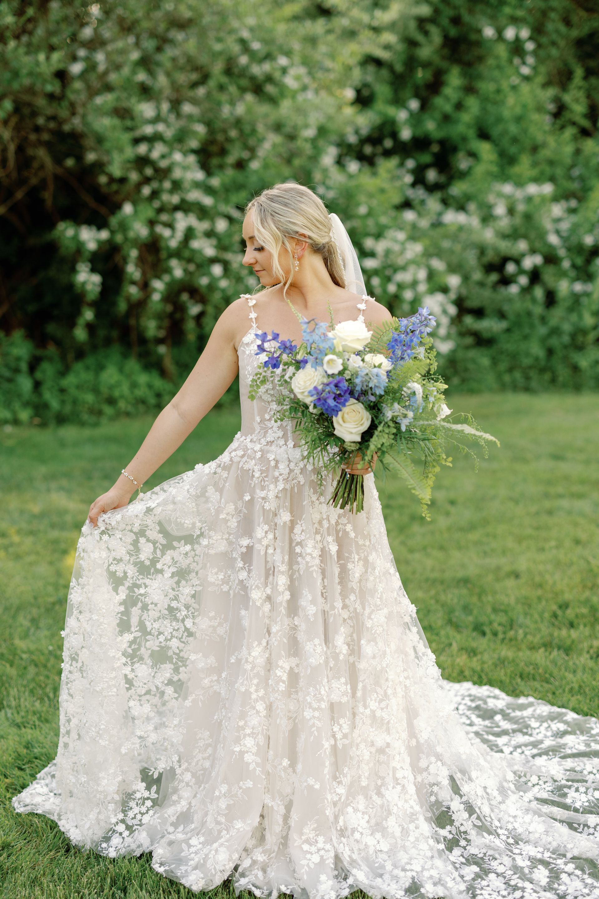 Bride in a lace wedding gown holding a blue-and-white bouquet in a garden