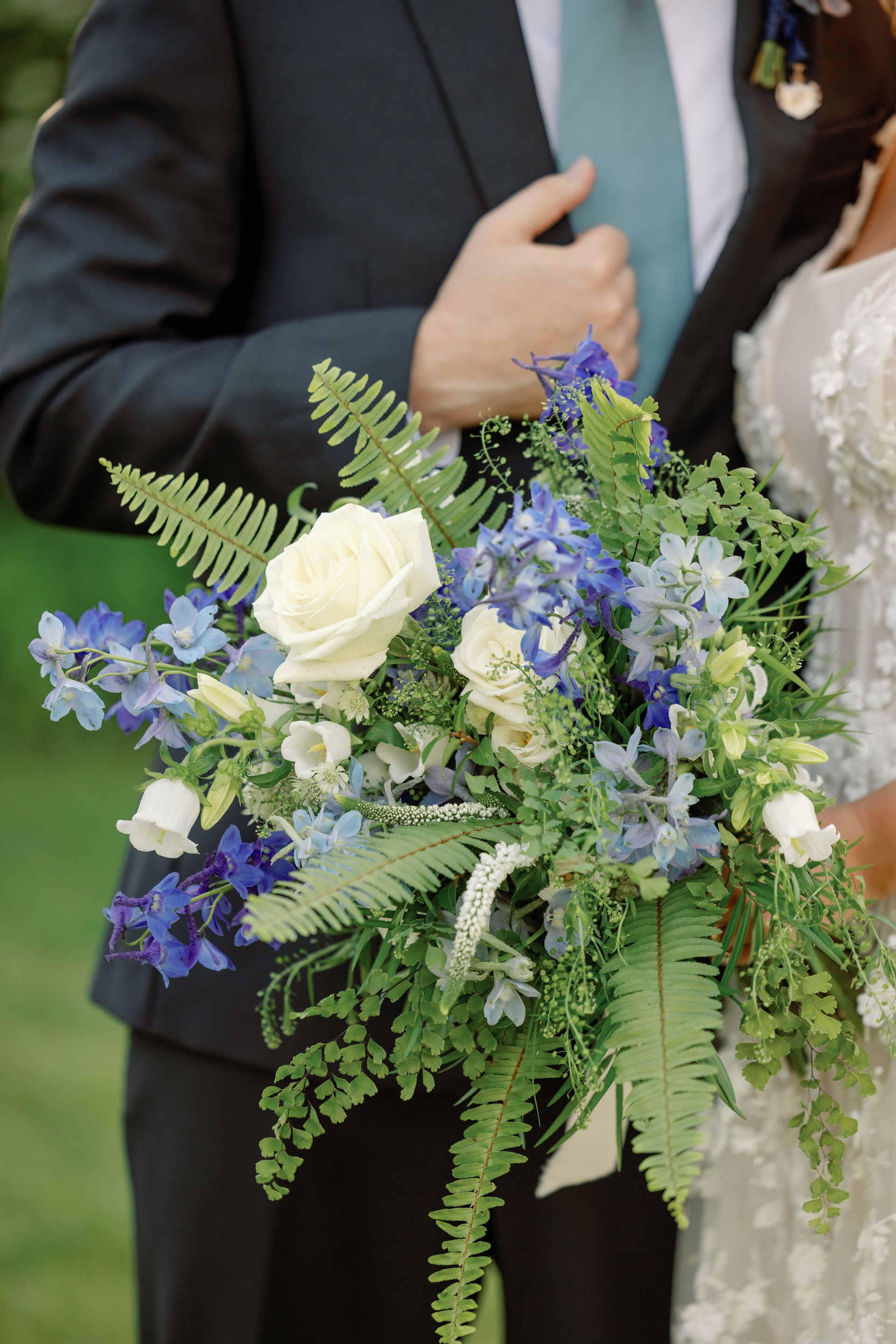 Wedding bouquet of white and blue flowers with ferns, held by someone in a dark suit and teal tie
