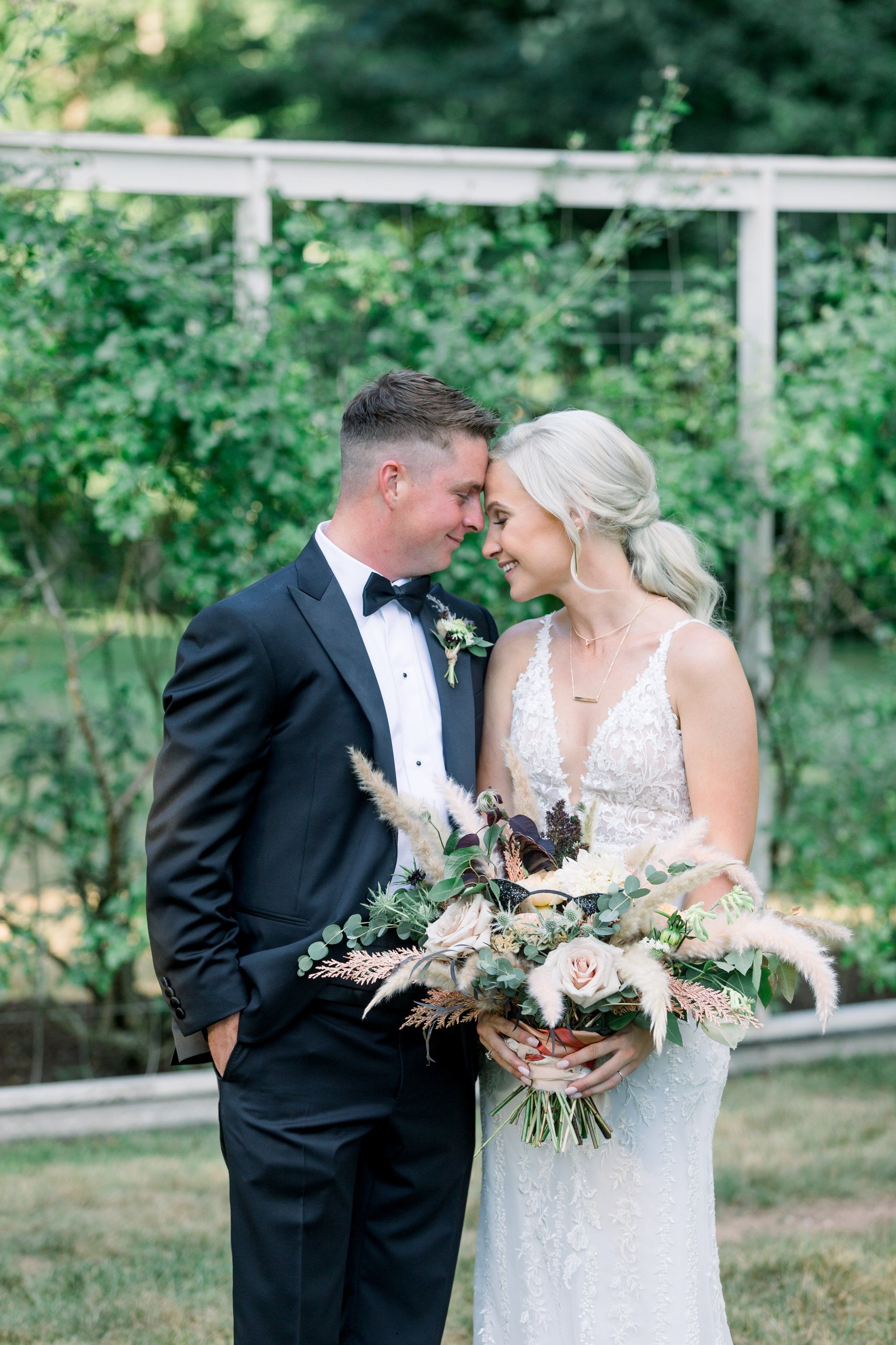 Bride and groom in black tuxedo and white lace dress sharing bouquets outdoors
