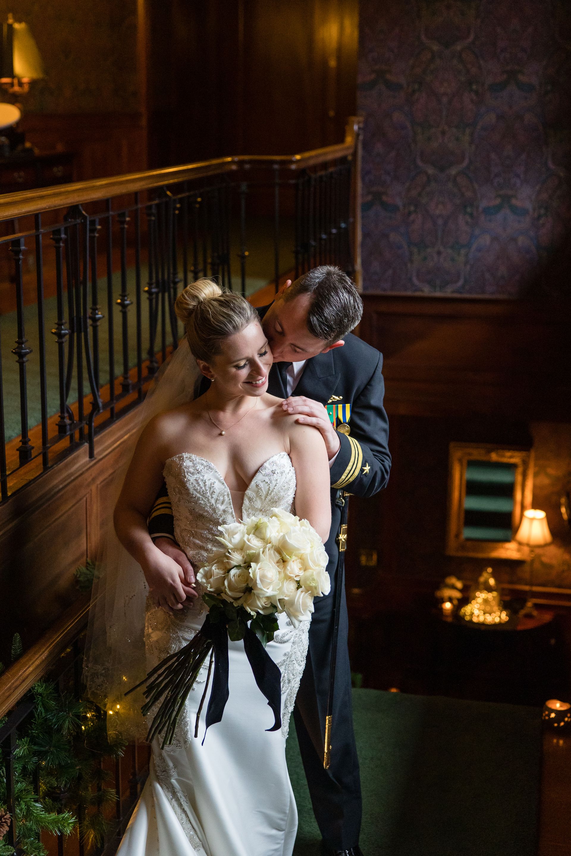 Bride and groom embracing on a staircase, bride holding a white bouquet, warmly lit interior