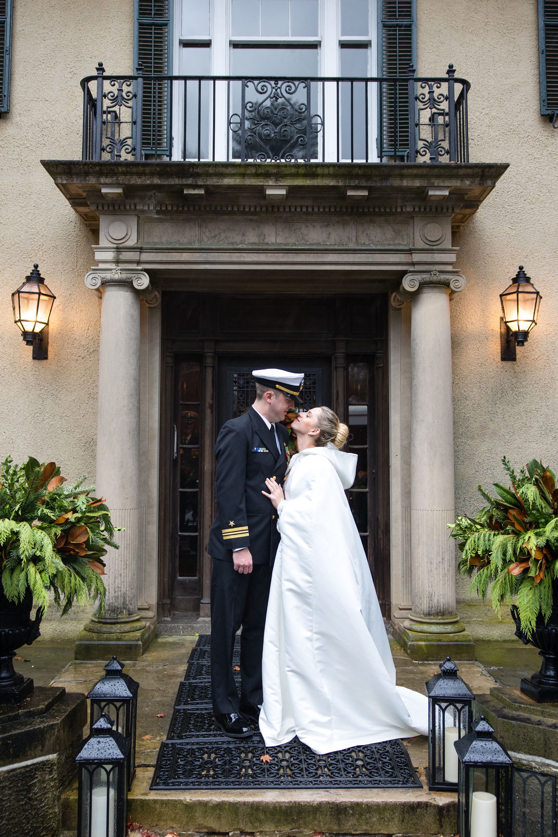 Bride and groom kissing outside a grand house entrance, framed by lanterns and potted plants.
