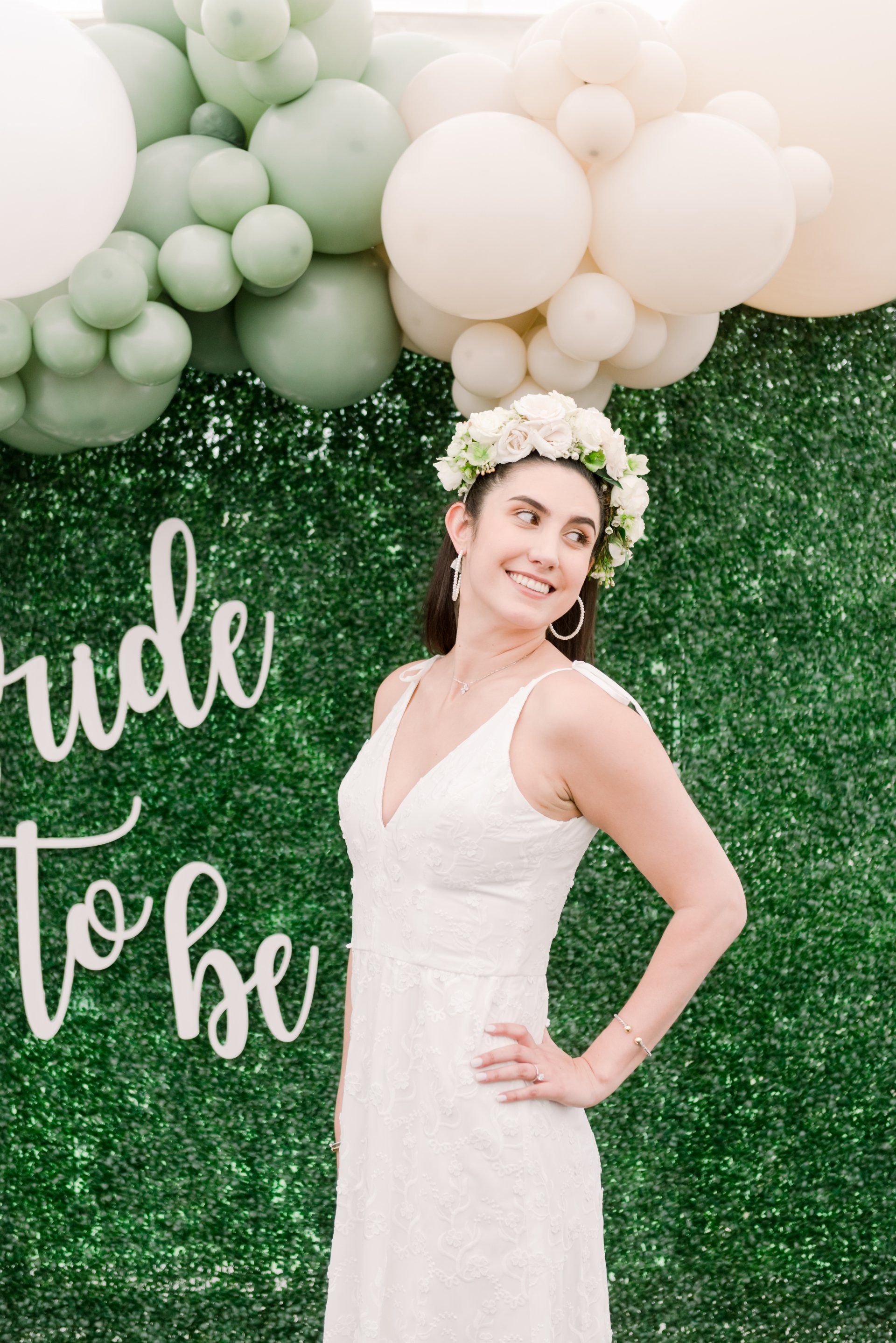 Woman in white dress posing at a bridal shower backdrop with green balloons and floral crown
