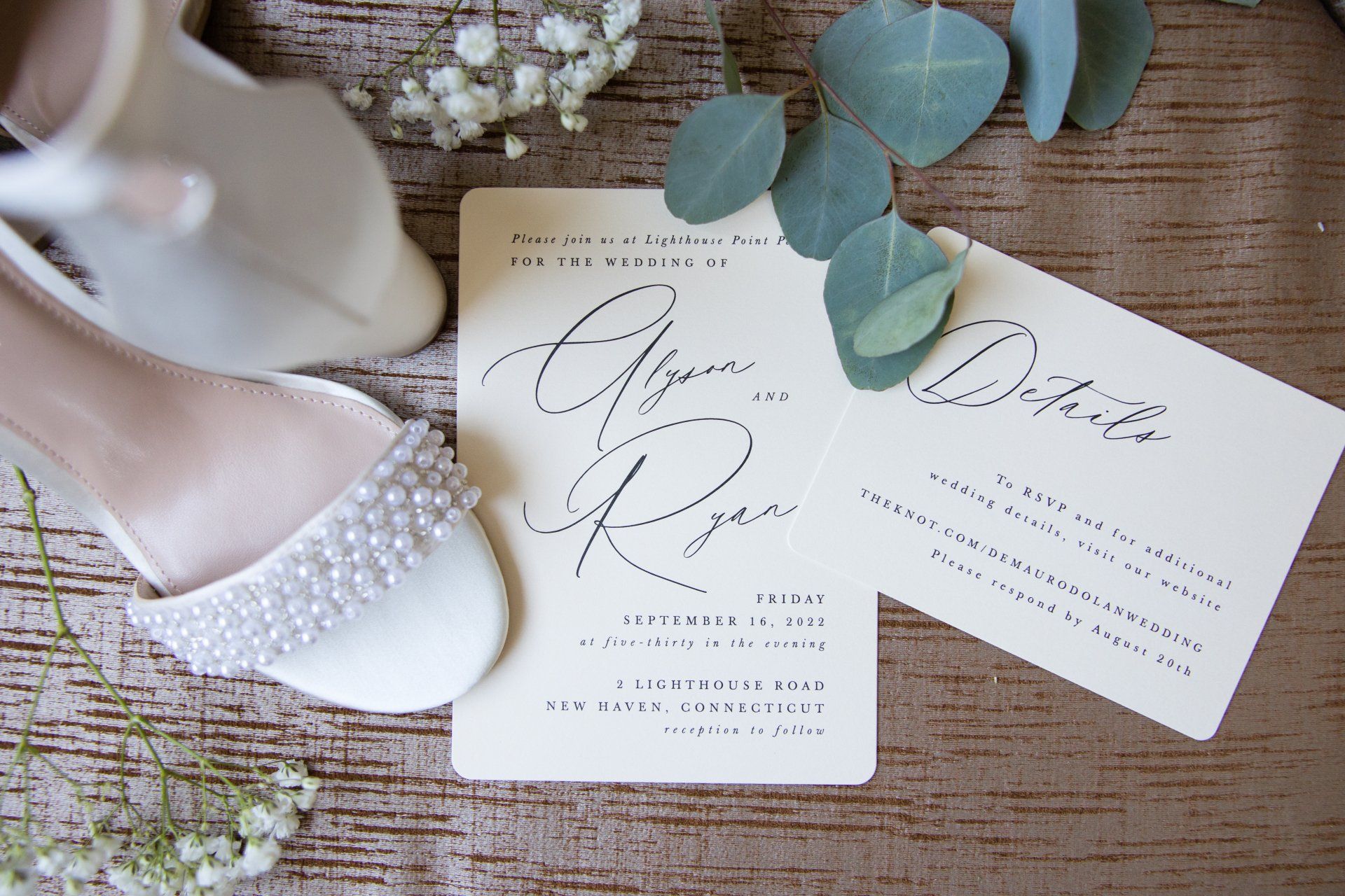 Pearl-detailed white shoes beside elegant wedding invitations and eucalyptus on burlap fabric