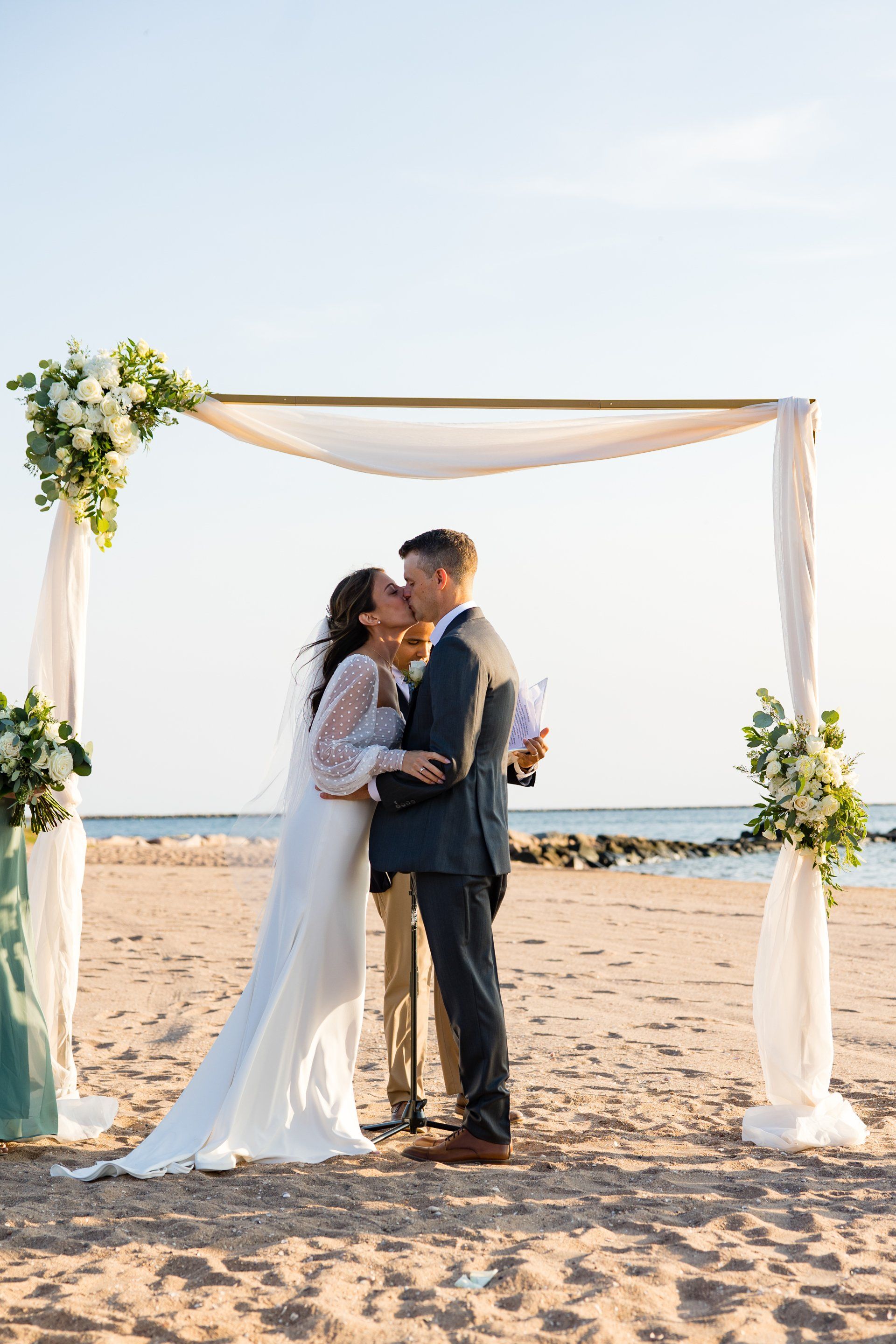 Couple embracing under a white wedding arch on a sandy beach at sunset