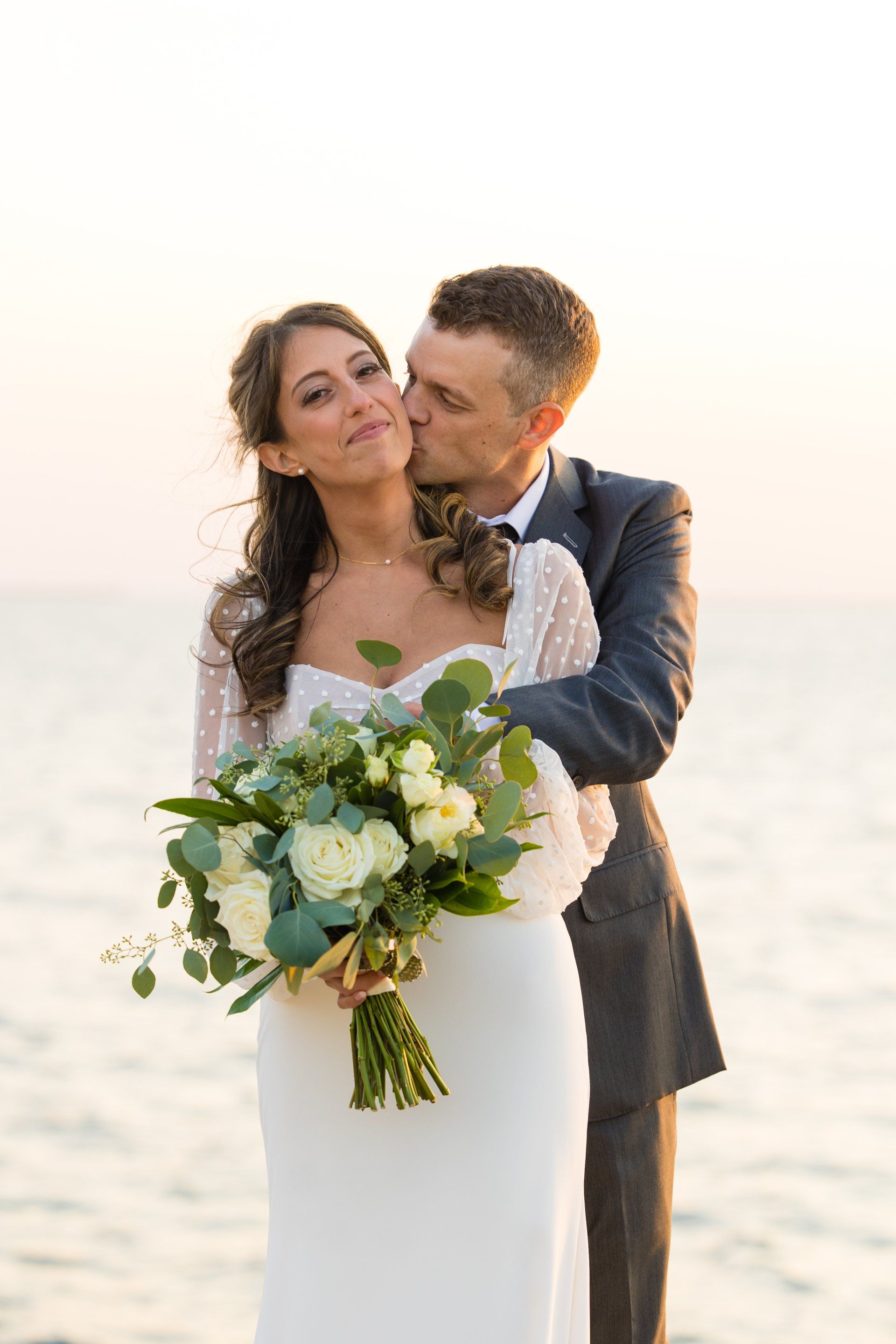 Couple embracing at the beach, bride holding a white-and-green bouquet at sunset
