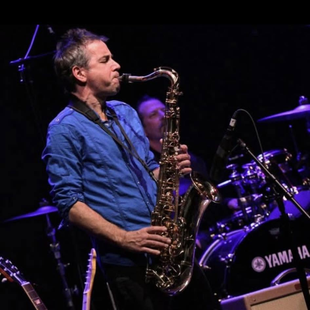 Musician in a blue shirt playing saxophone on a dimly lit stage with drums behind him