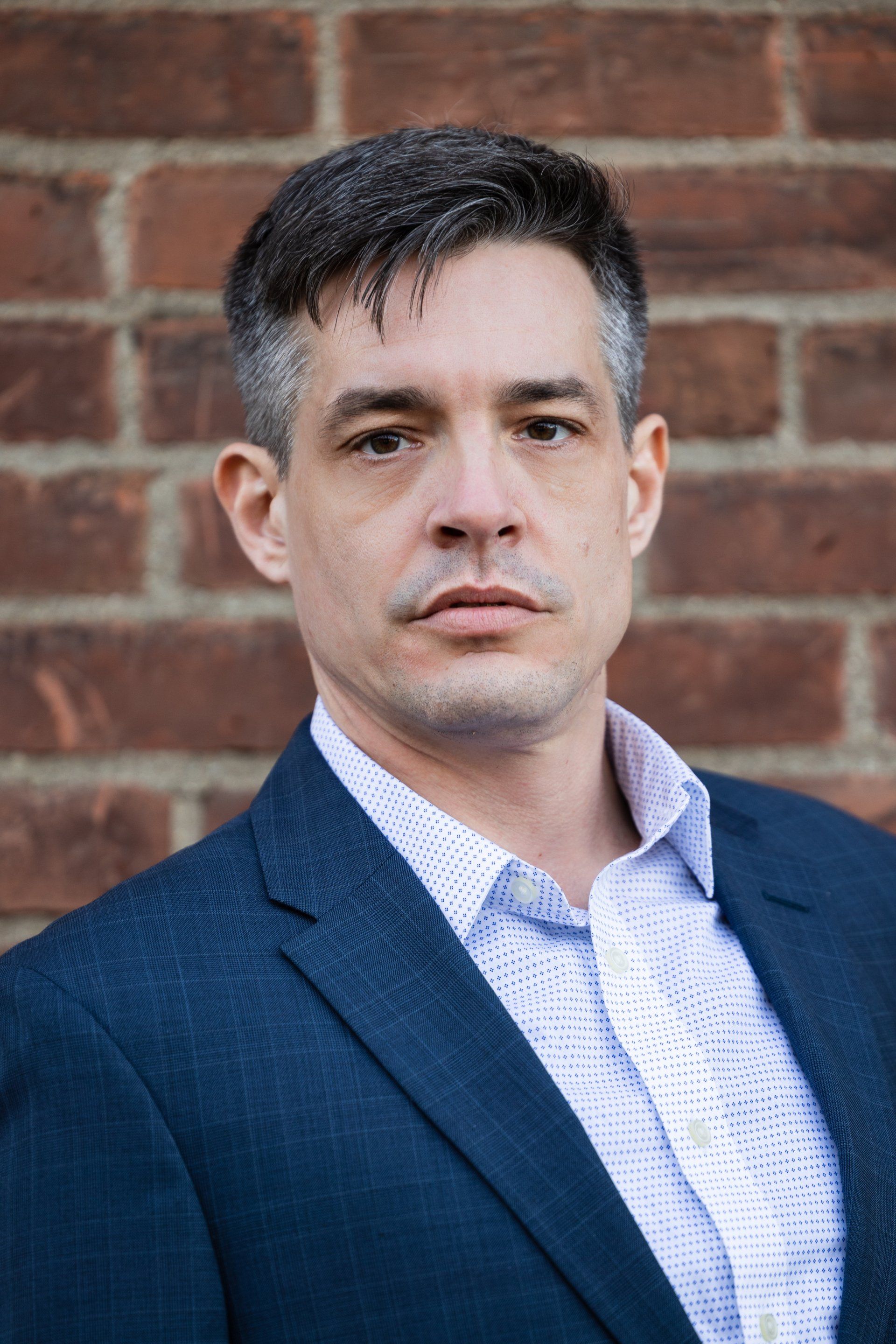 Man in a blue blazer and white shirt standing against a brick wall, looking at the camera.