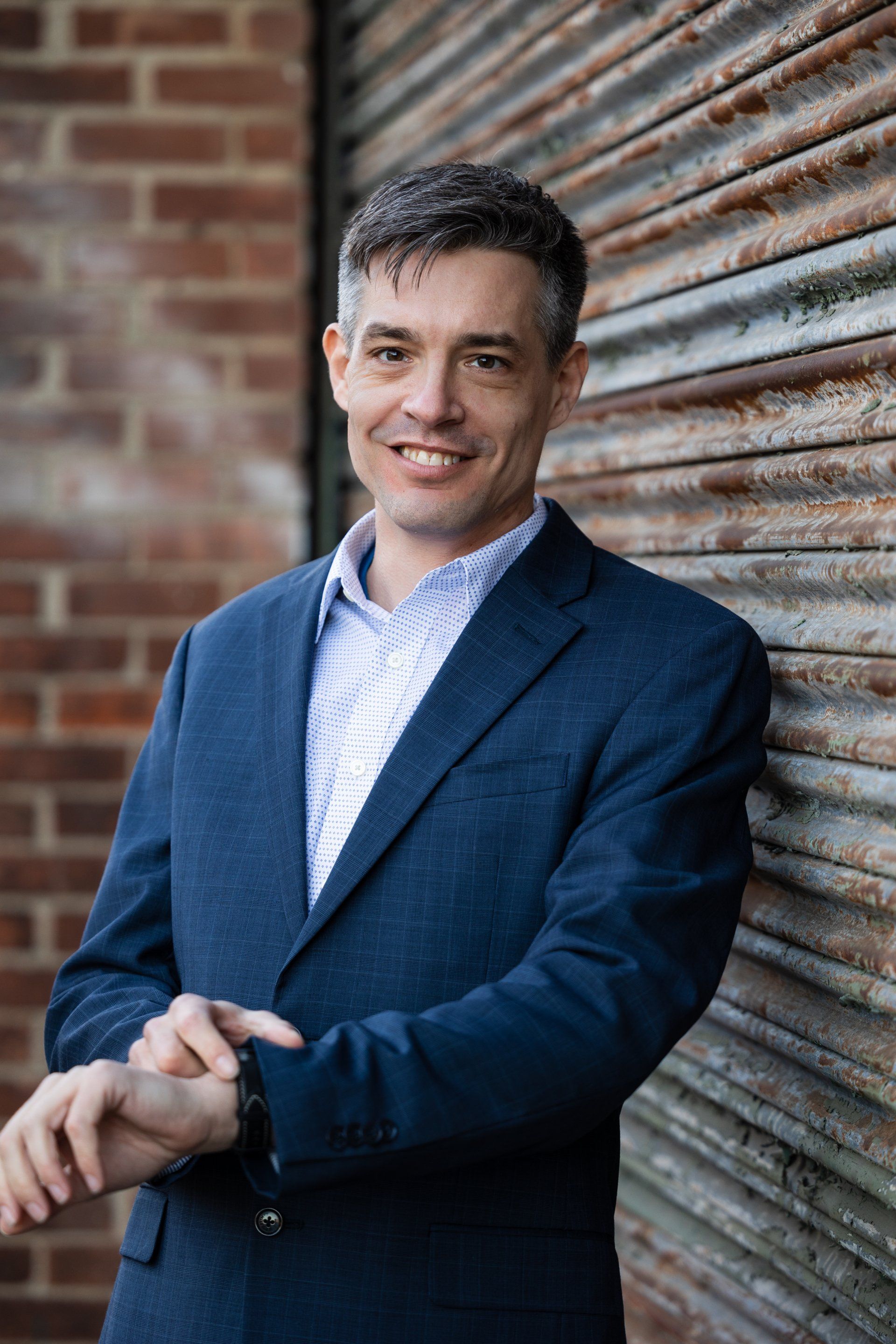Man in a navy blazer leaning against a brick wall and corrugated metal door, smiling at the camera