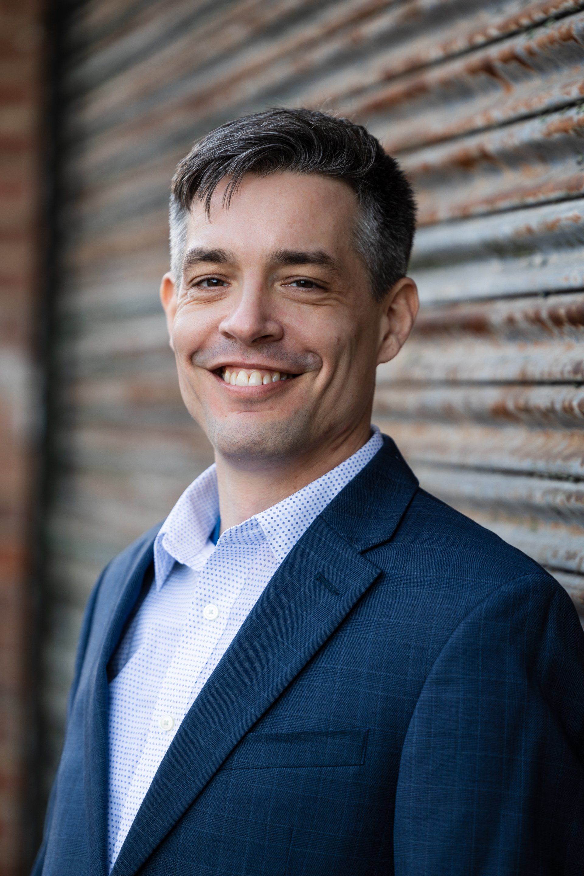 Smiling man in a blue blazer standing against a weathered wooden wall.