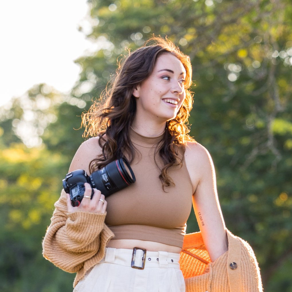 Woman holding a camera outdoors in sunlight, wearing a beige top and white pants, smiling