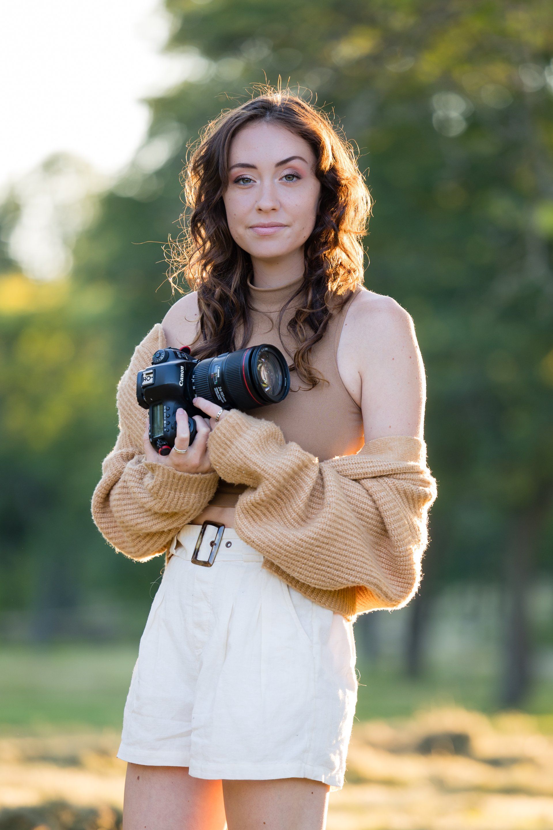 Woman holding a camera outdoors in a sunlit park, wearing a tan sweater and white skirt.