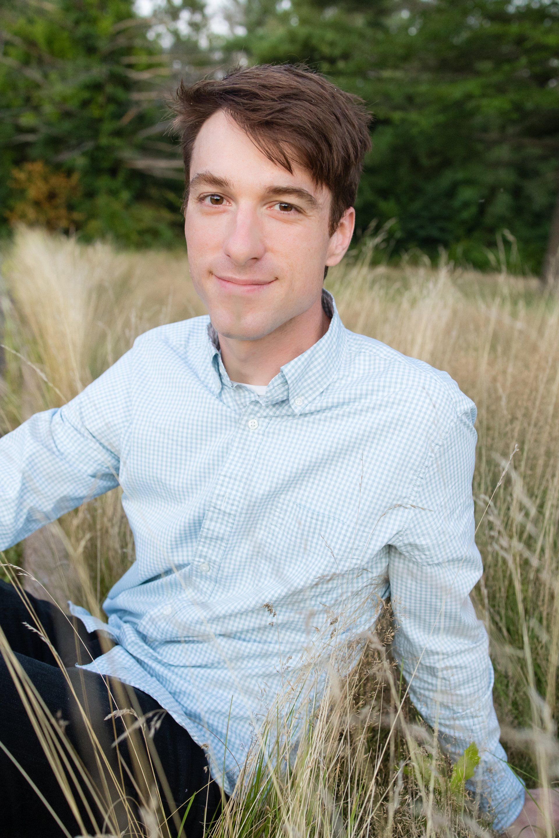 Person sitting in tall grass outdoors, wearing a light blue shirt and smiling at the camera