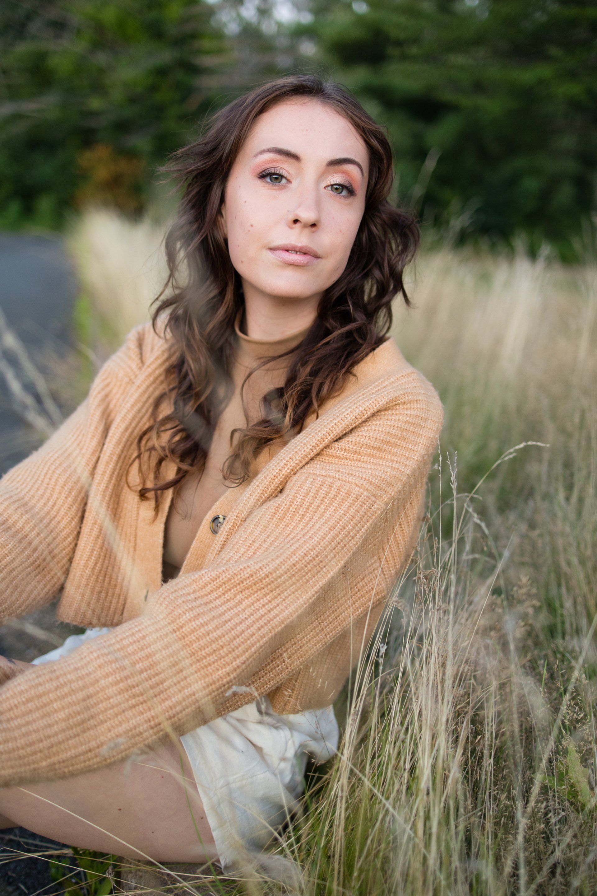 Woman in a tan sweater sitting in tall grass, looking at the camera.