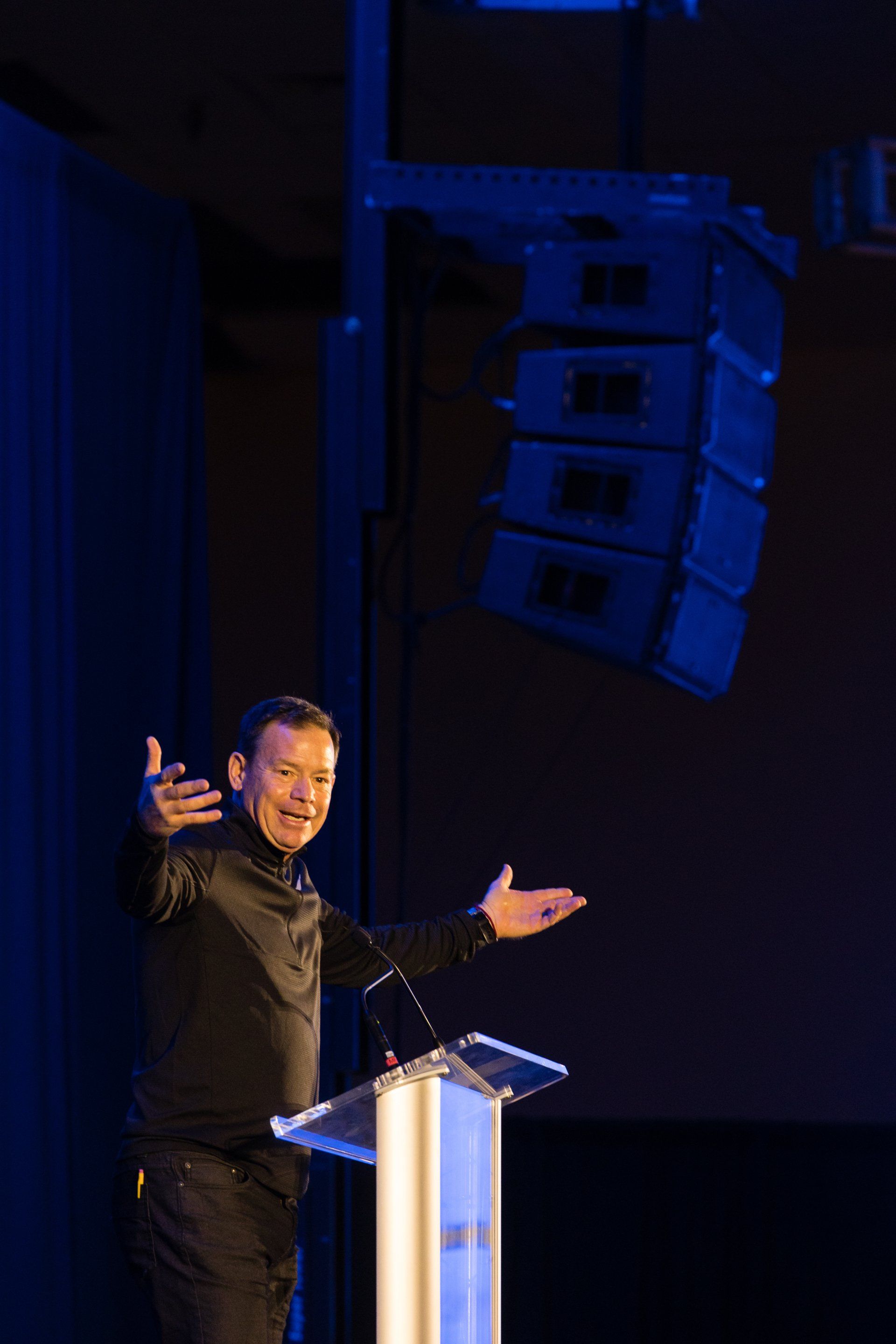 Man speaking at a podium with arms outstretched on a dark stage, blue-lit speaker array behind him