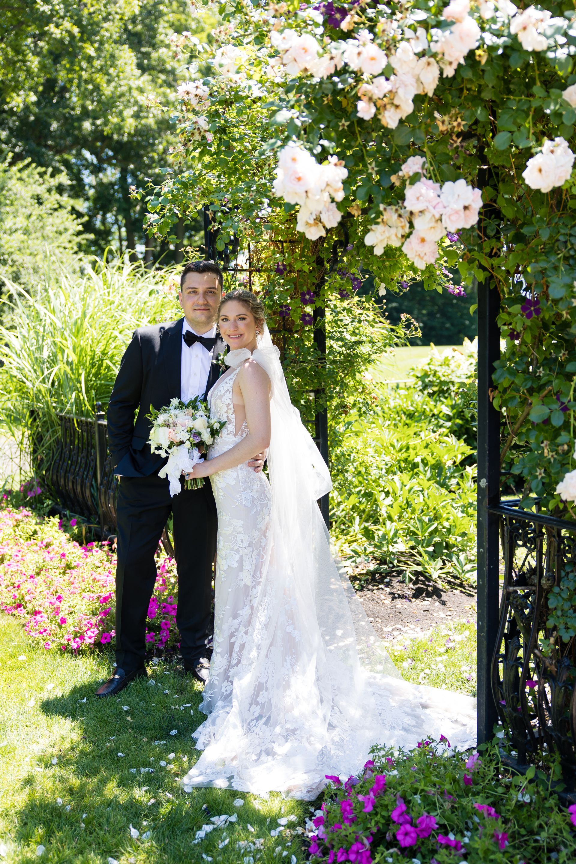 Bride and groom under a blooming arch in a garden, holding a bouquet and smiling.