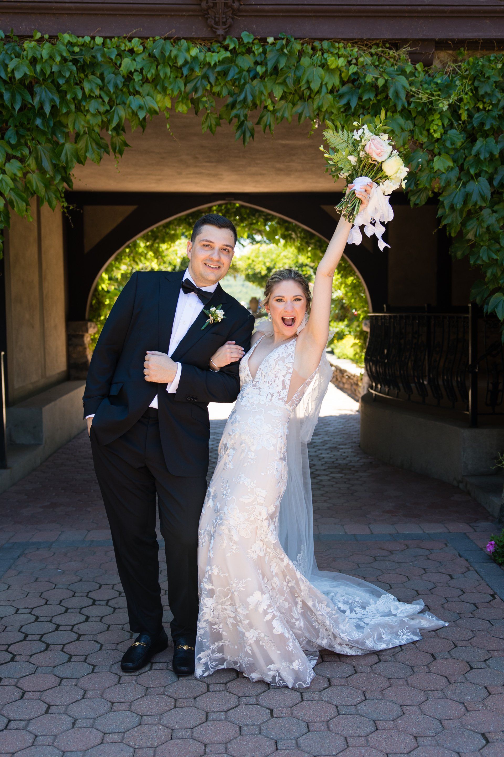 Bride and groom pose under ivy, the bride holding a bouquet and raising it overhead