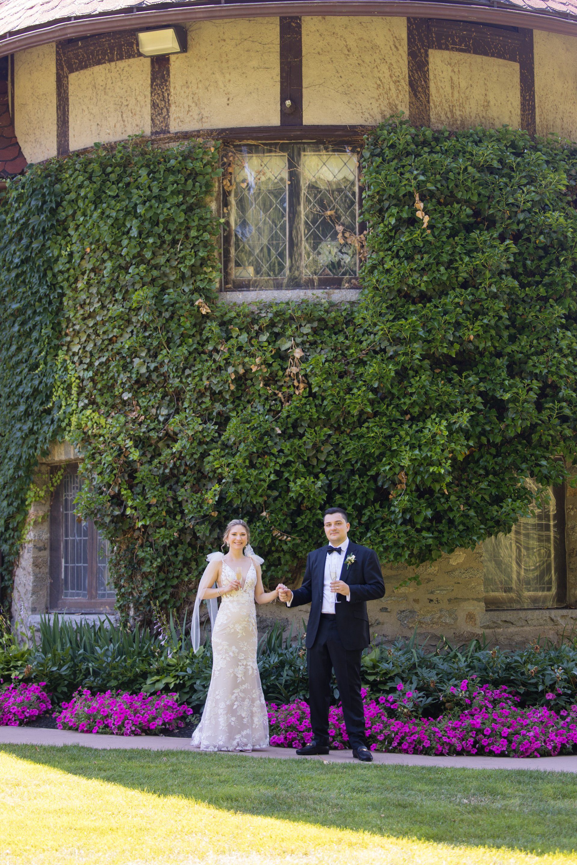 Bride and groom standing in front of a vine-covered building with pink flowers and green lawn