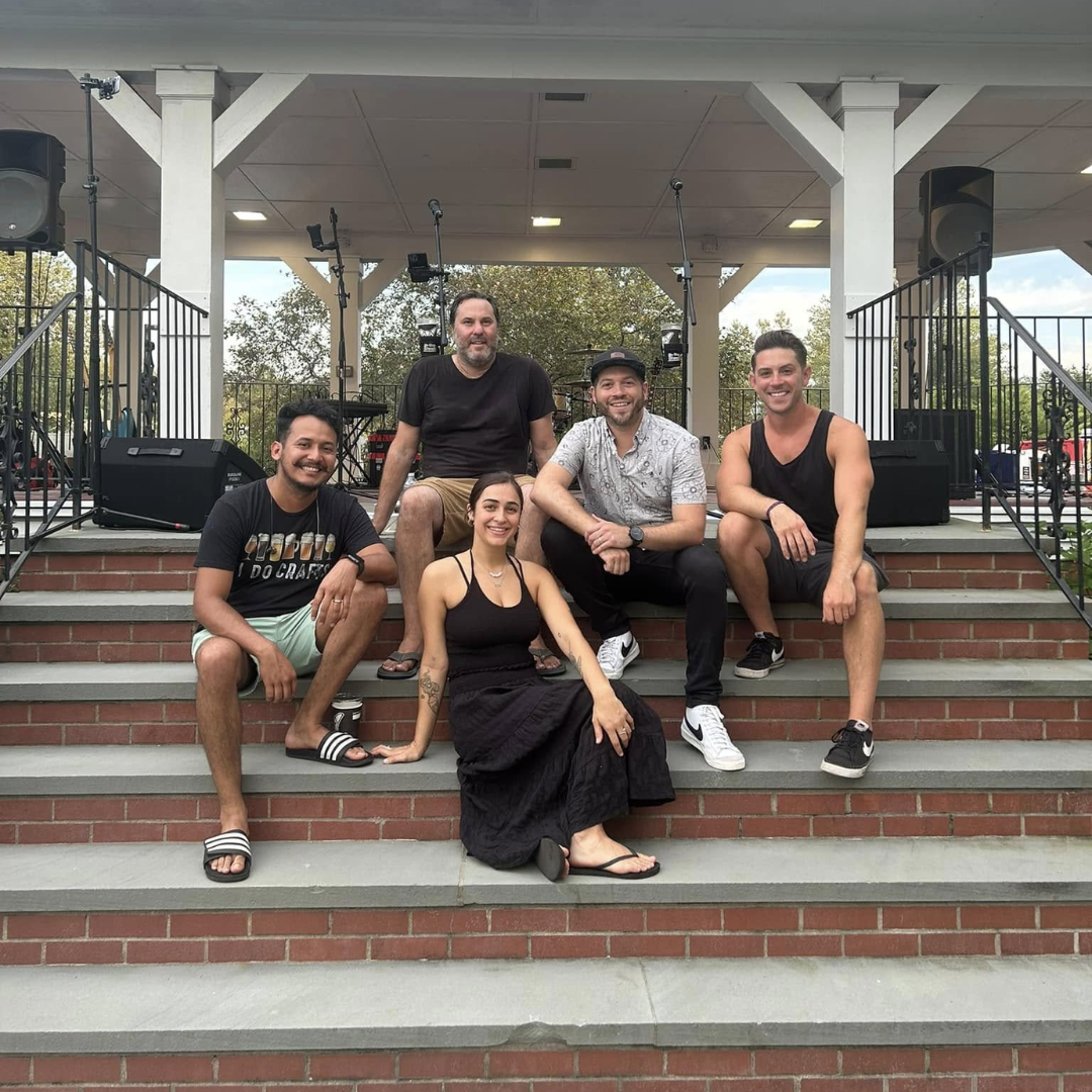 Five people sit and pose on outdoor pavilion steps, smiling in casual clothes.