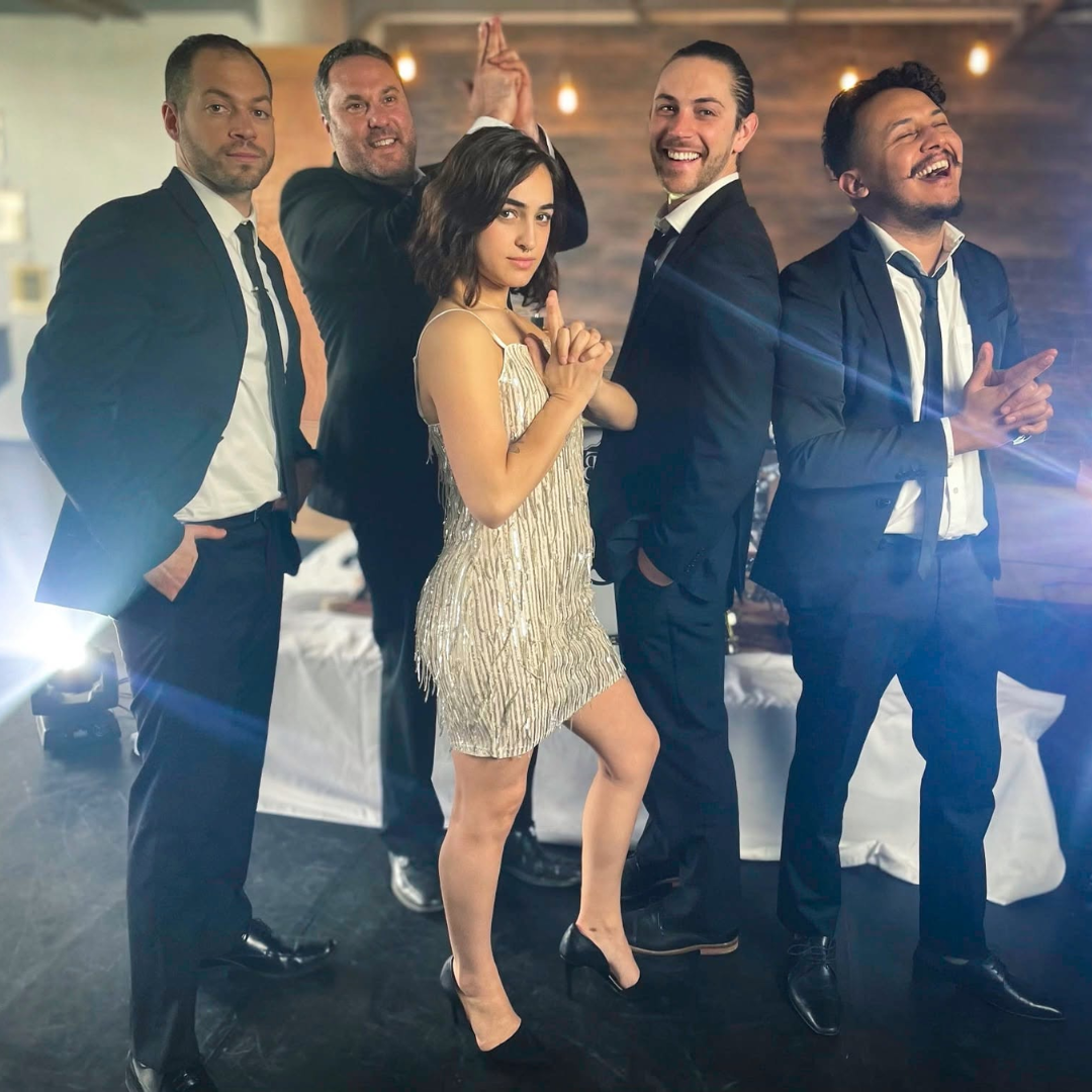 Five people in formal wear posing and smiling at a party under warm lights