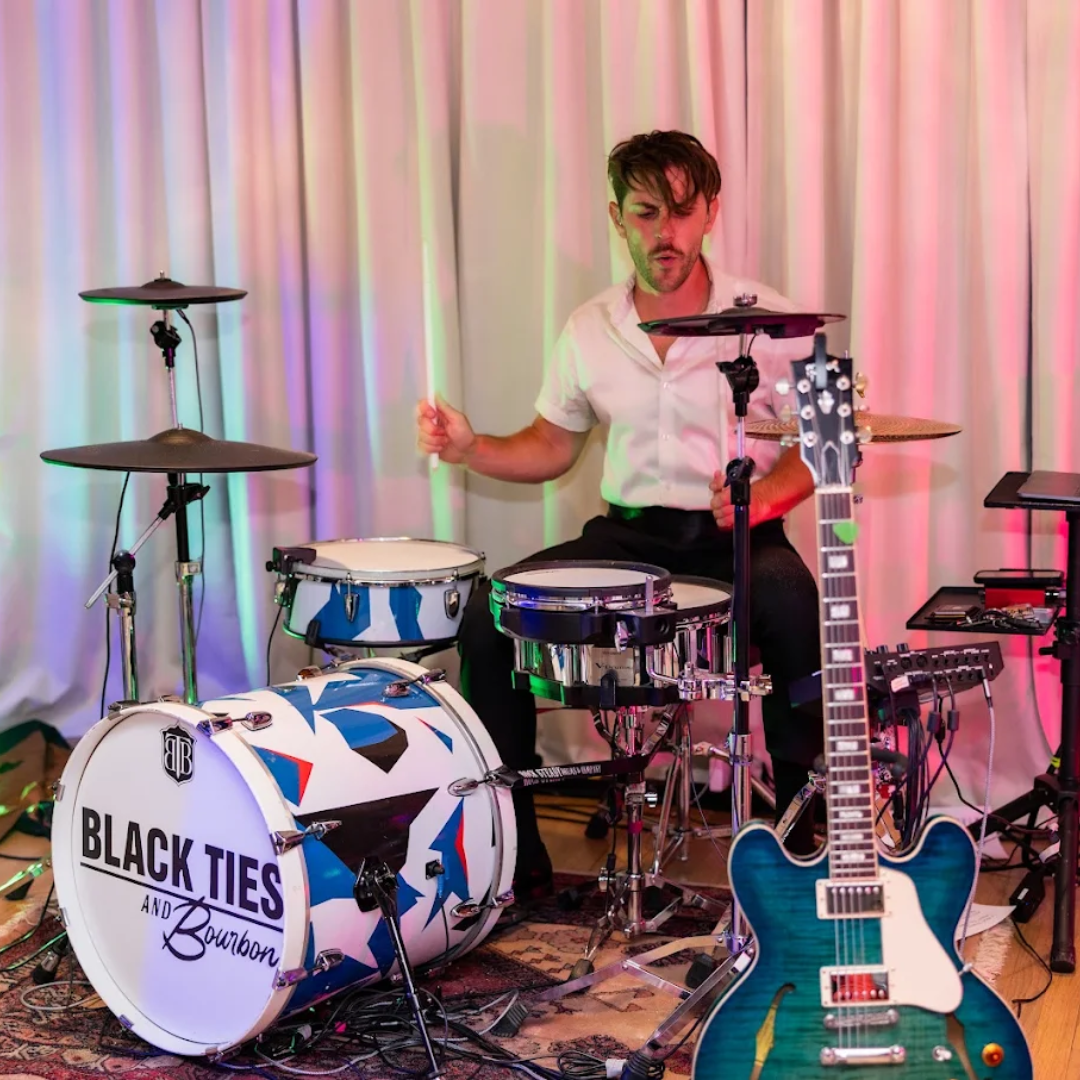 Drummer behind a blue-and-white kit with a blue guitar in front, standing against pink curtains
