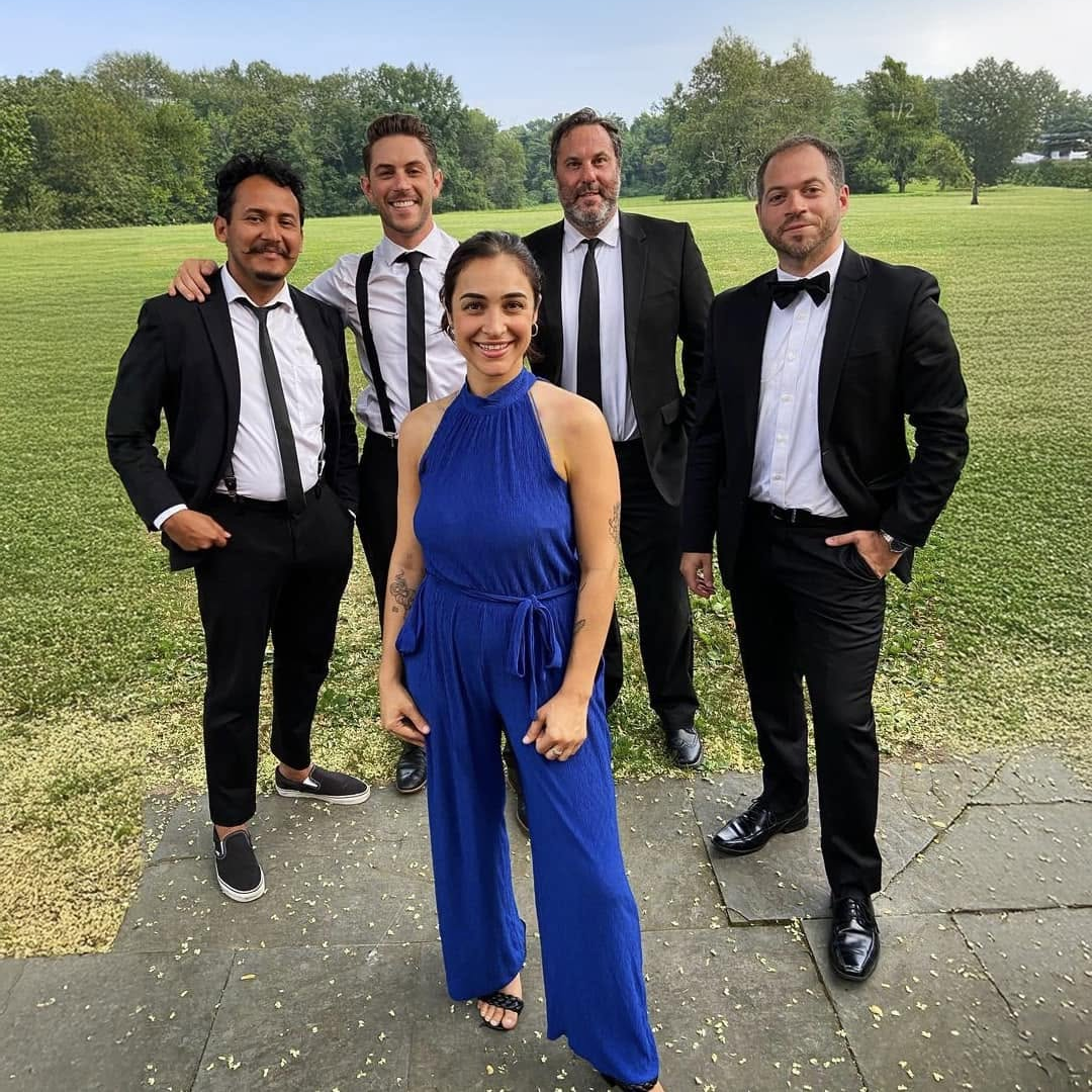 Five people dressed formally standing outdoors on a sidewalk in a park-like setting