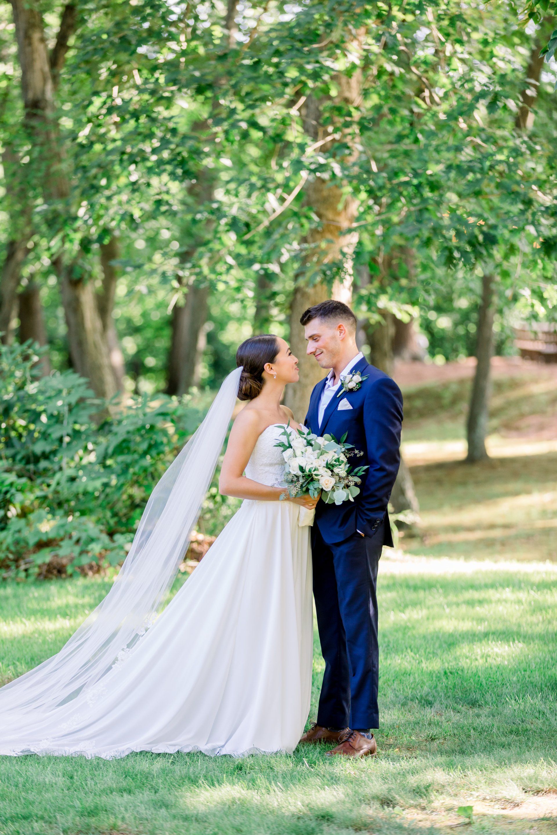 Bride and groom standing together in a leafy park, holding bouquets and facing each other under trees
