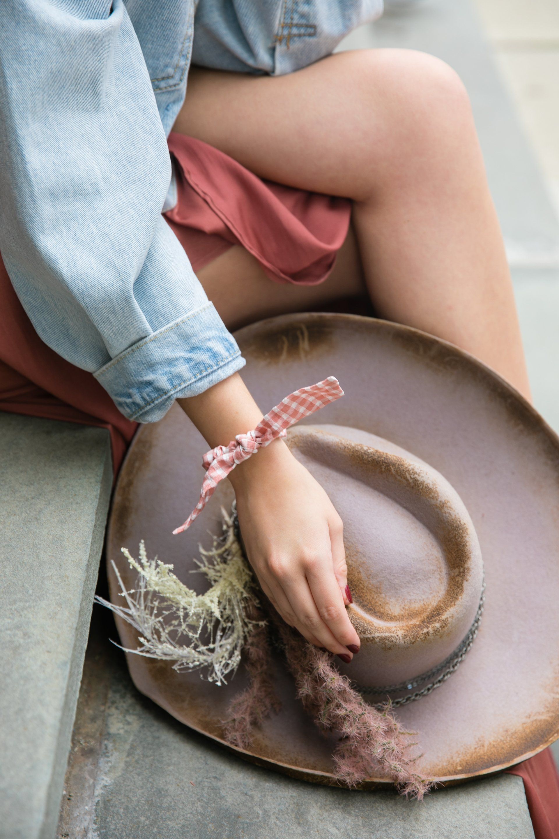 Person holding a brown straw hat with dried flowers and a pink ribbon while sitting in a denim shirt and pink skirt