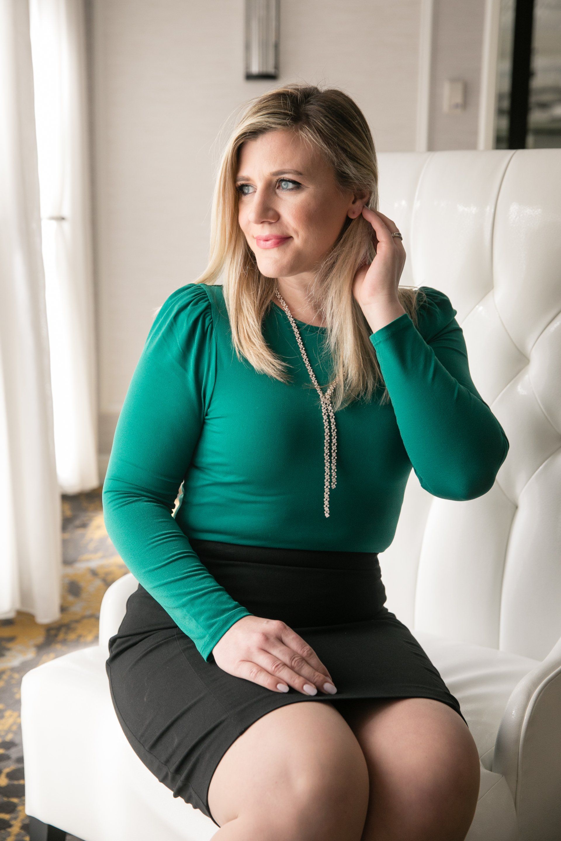 Woman in a green top sitting on a white chair, adjusting her hair near a bright window.