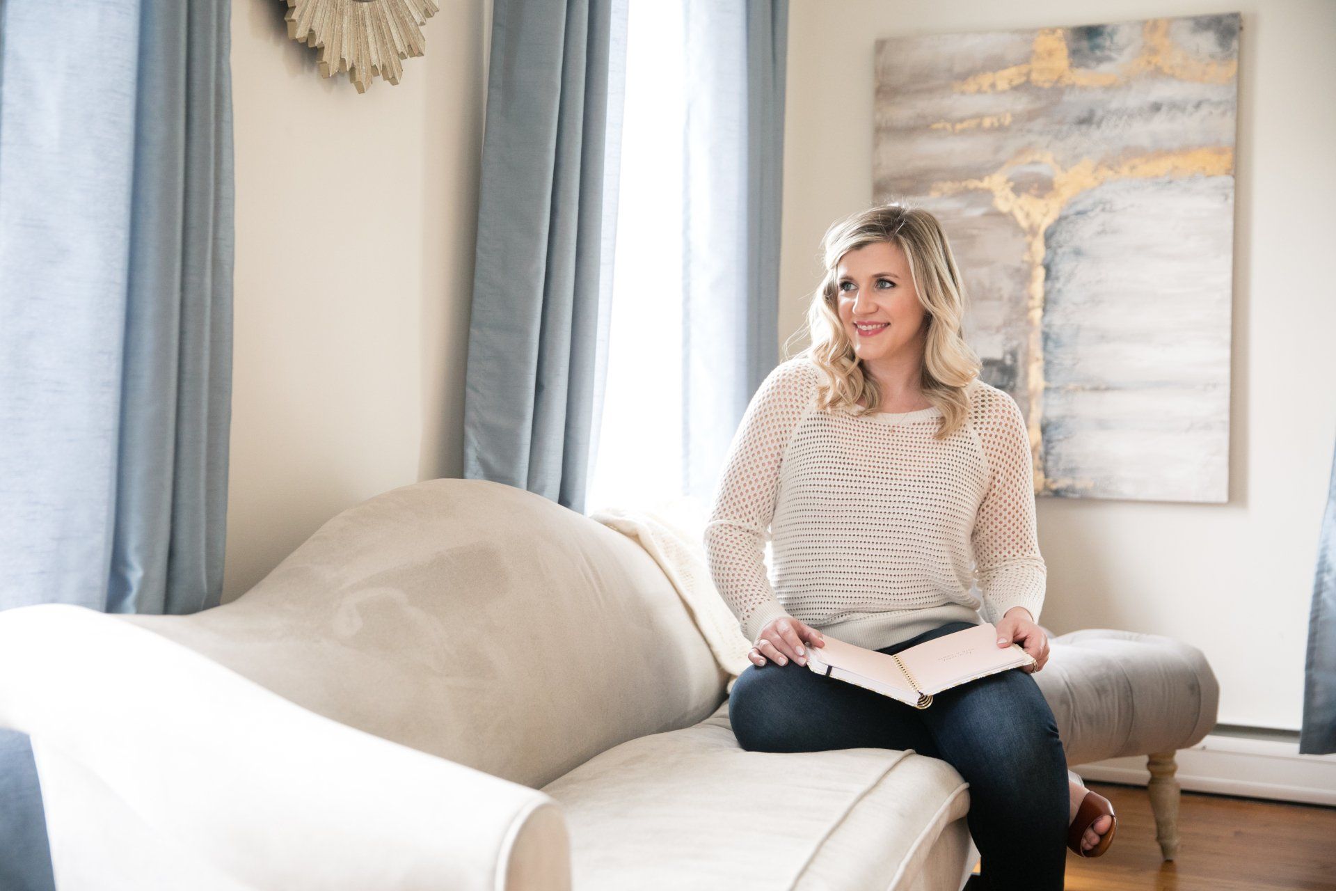 Woman seated on a beige sofa in a bright living room, holding a notebook and smiling toward the window.