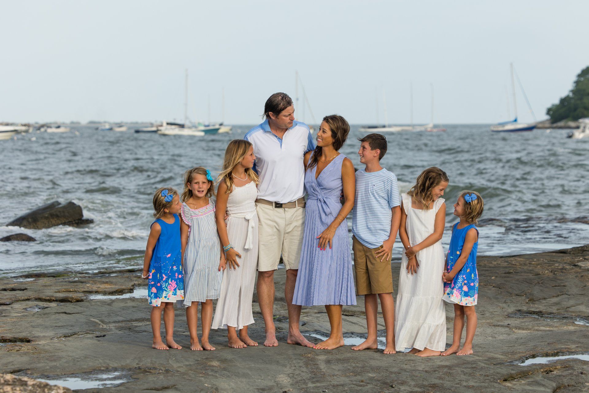 Group of people standing on a rocky beach by the water, with sailboats in the background.
