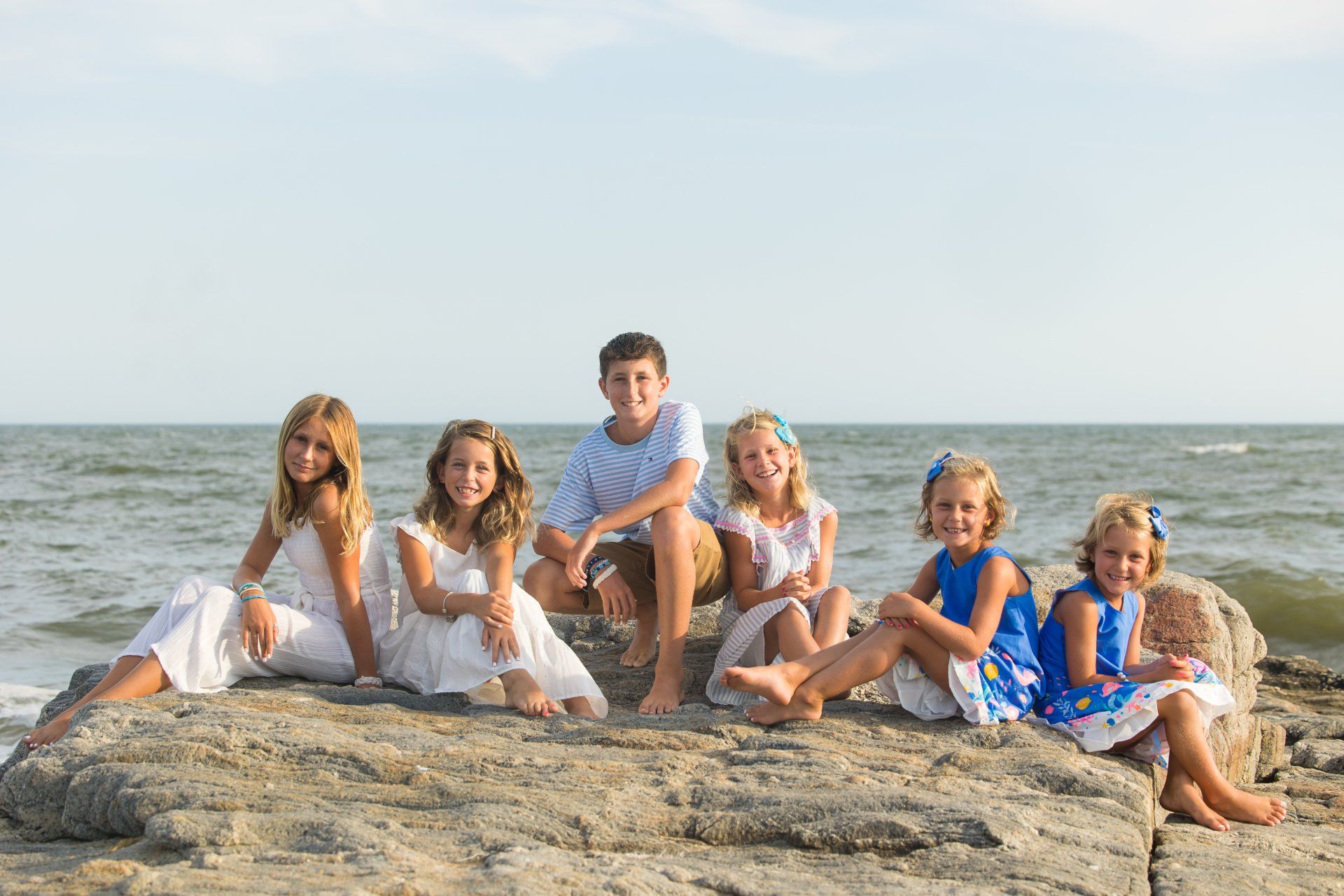 Group of six sitting on seaside rocks, smiling at the ocean under a bright sky.