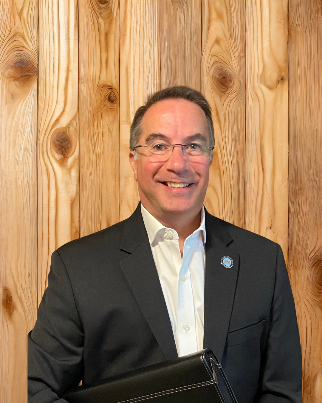 Smiling man in a dark suit holding a black portfolio, standing against a wooden wall.