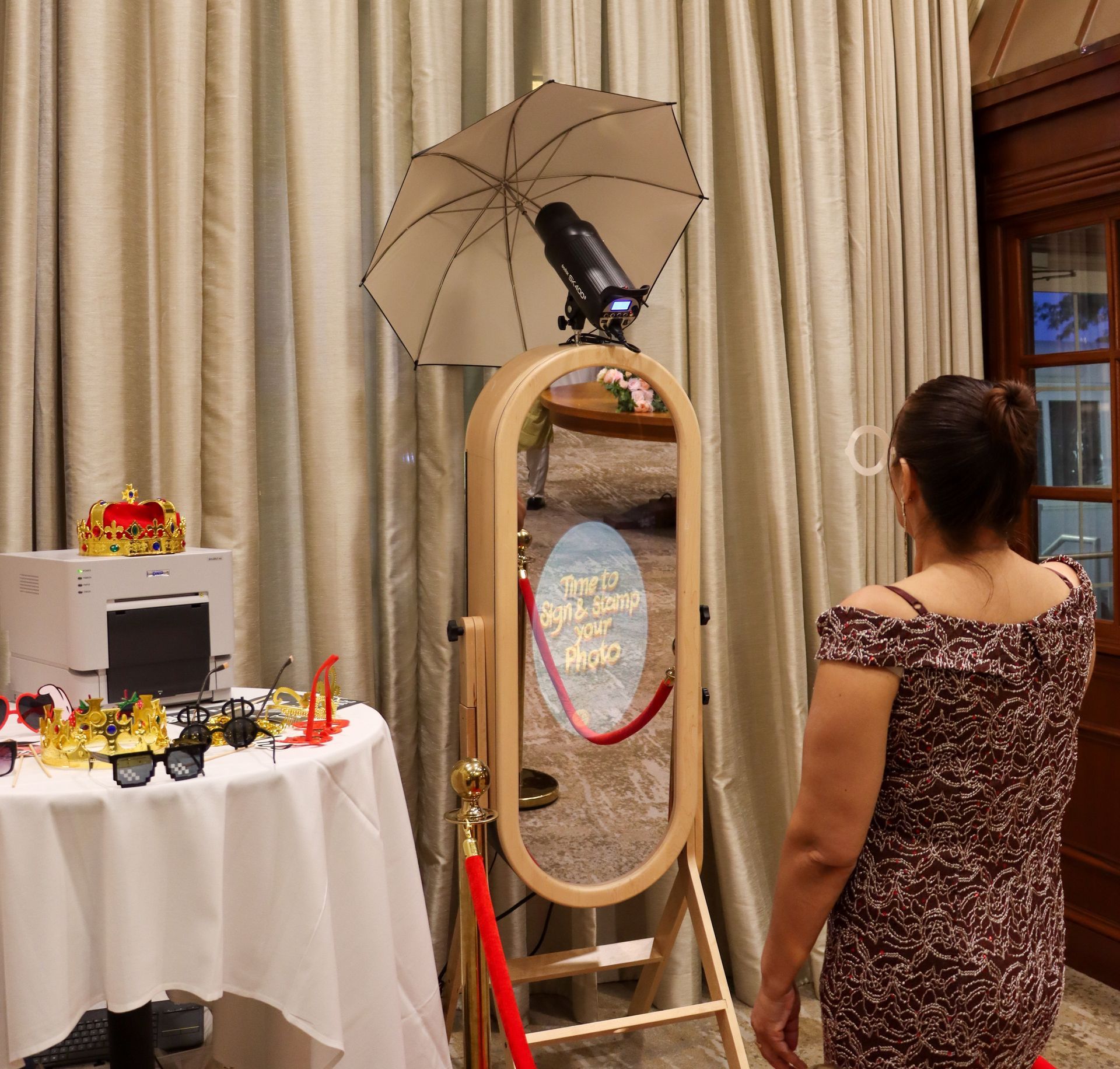Woman looks into a standing mirror with a camera and umbrella attached in a curtained room.