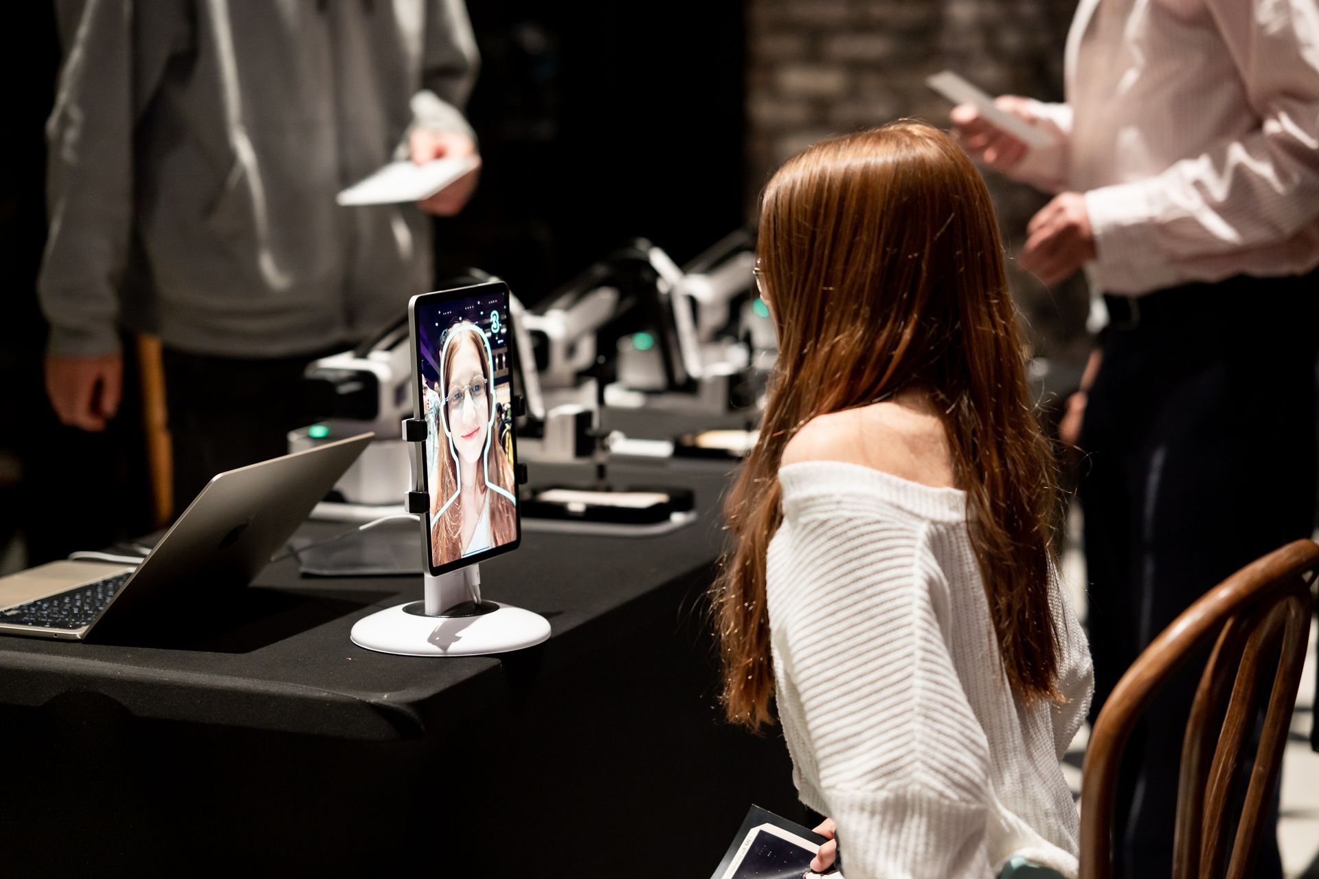Person in white sweater watches a tablet on a table in a dim event space, with others nearby