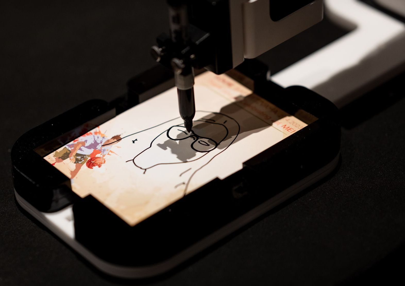 A pen plotter drawing a cartoon character on paper with black ink and colorful paint accents.