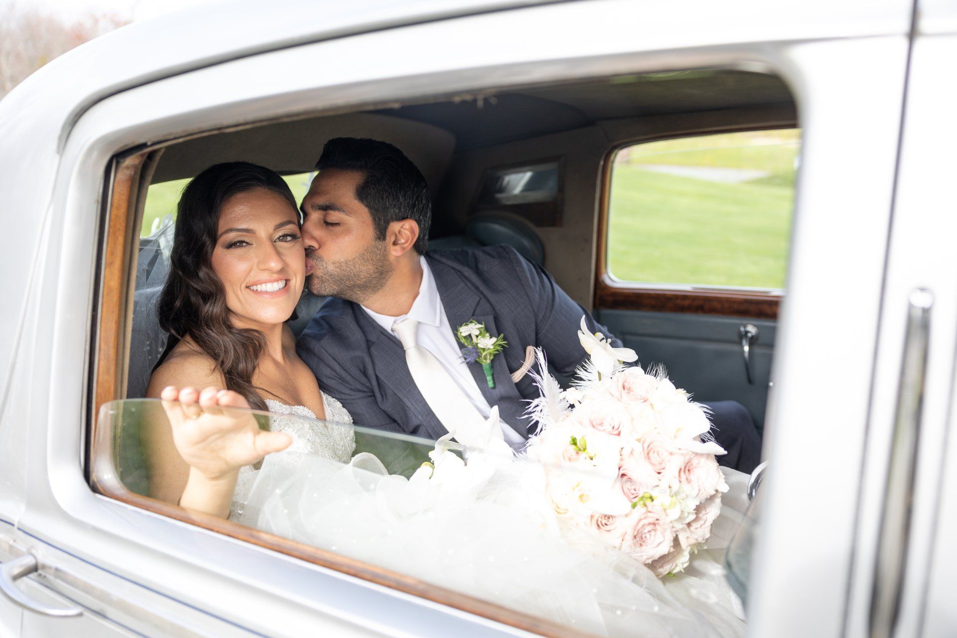 Bride and groom smiling in a car, with the groom kissing her cheek and a bouquet in the foreground