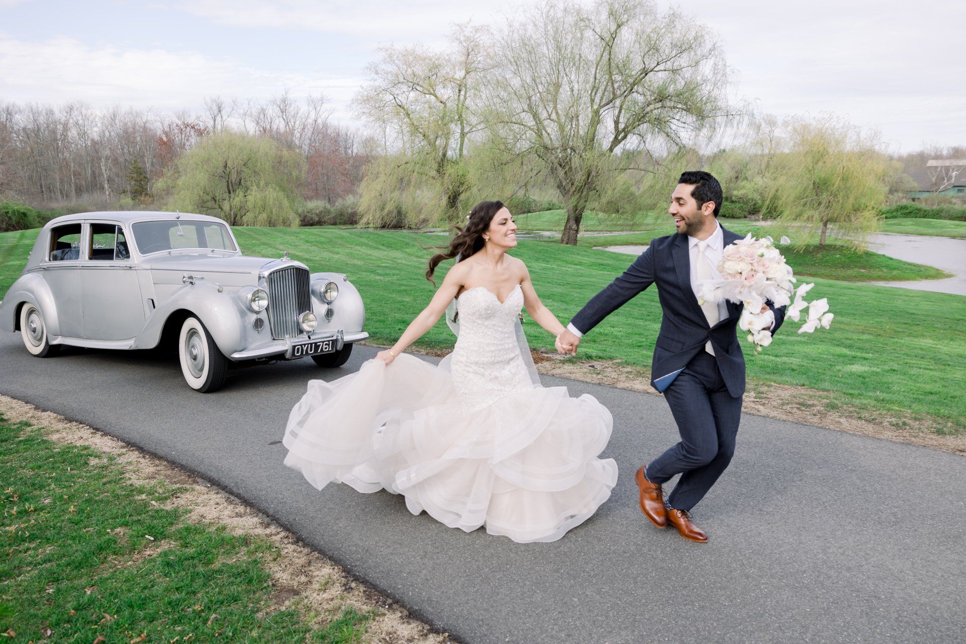 Bride and groom running hand in hand beside a vintage silver car on a park road
