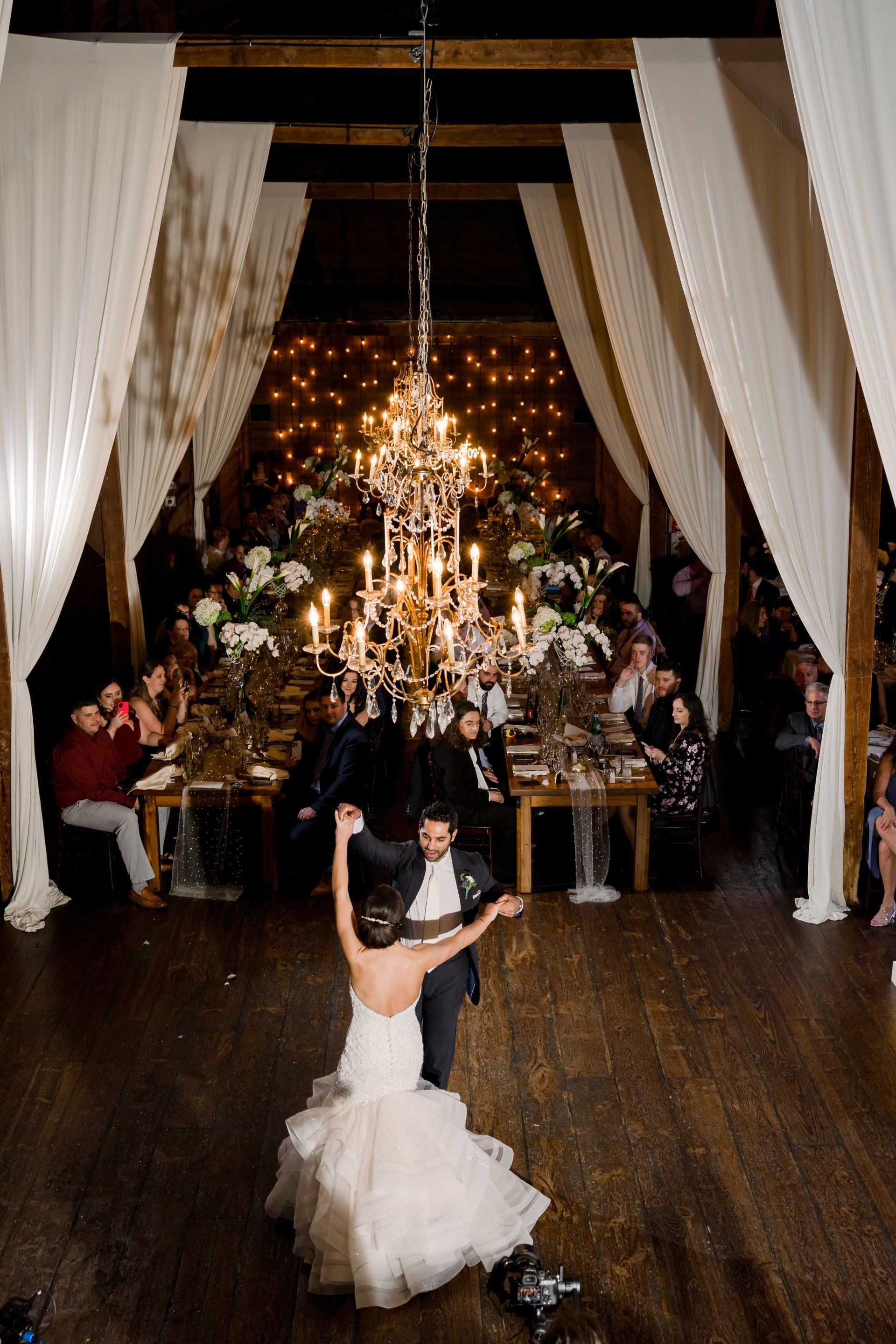 Bride and groom dancing in a candlelit hall beneath a chandelier, with guests seated on either side.