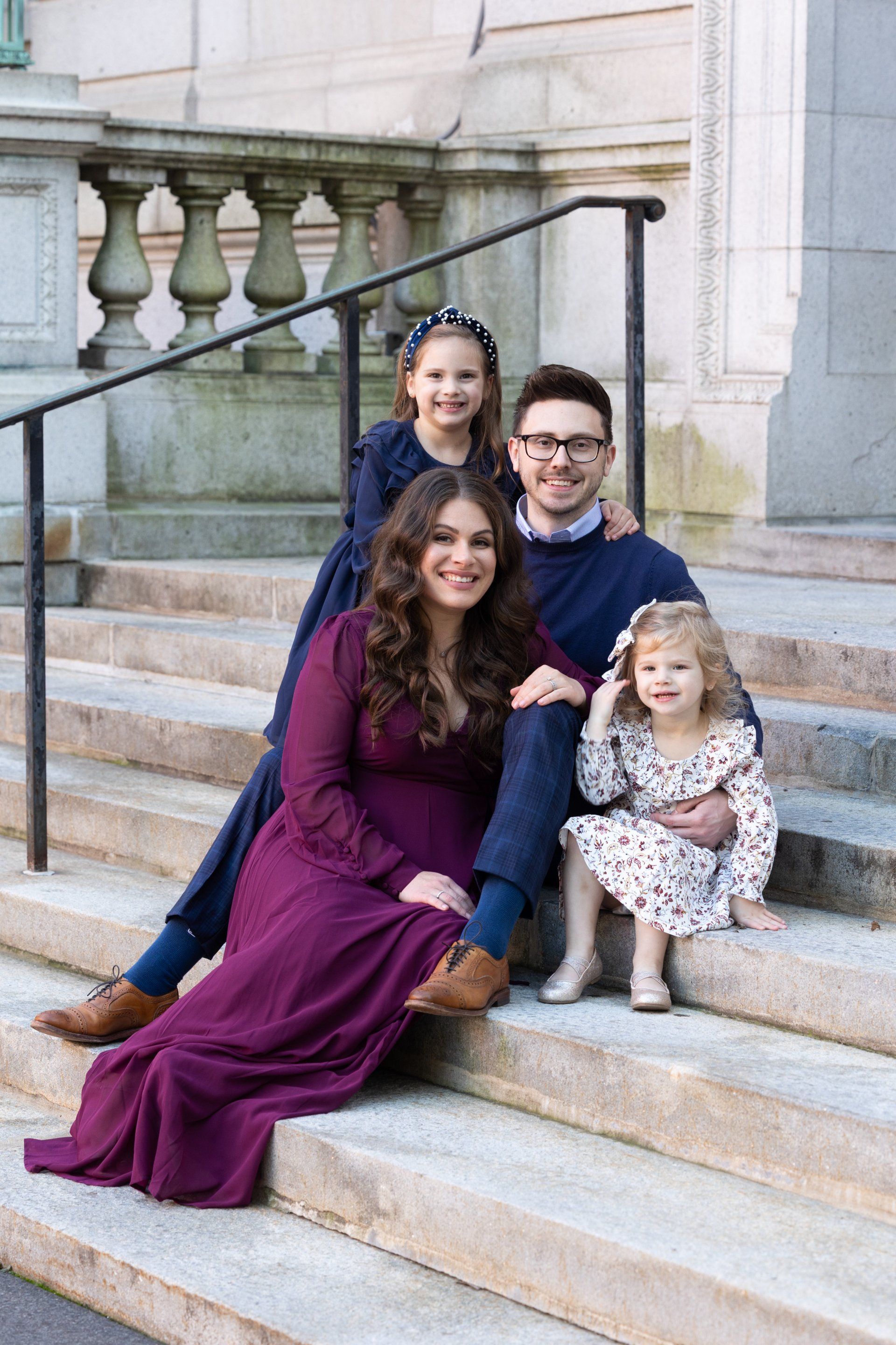 Family of four seated on stone steps, smiling in front of a building