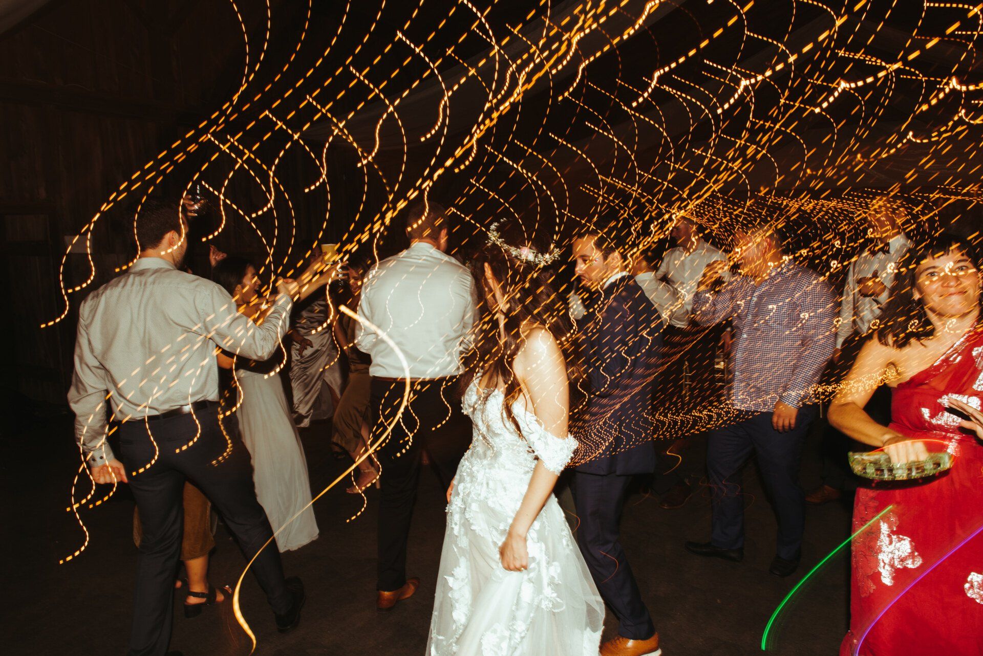 People dancing at a party with golden light trails and a woman in a white dress in the center