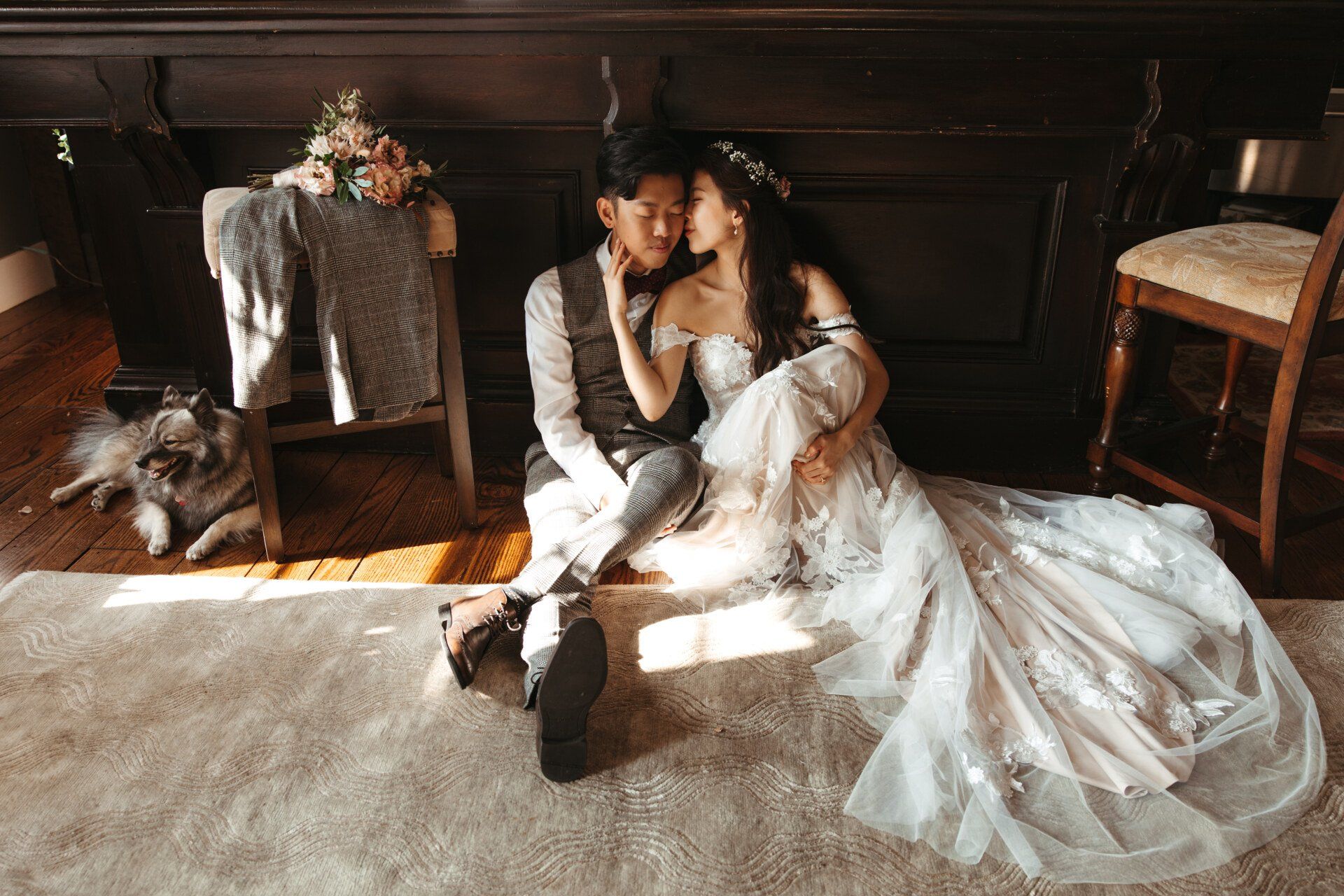 Couple in white wedding attire seated on the floor by a dark fireplace in a sunlit room