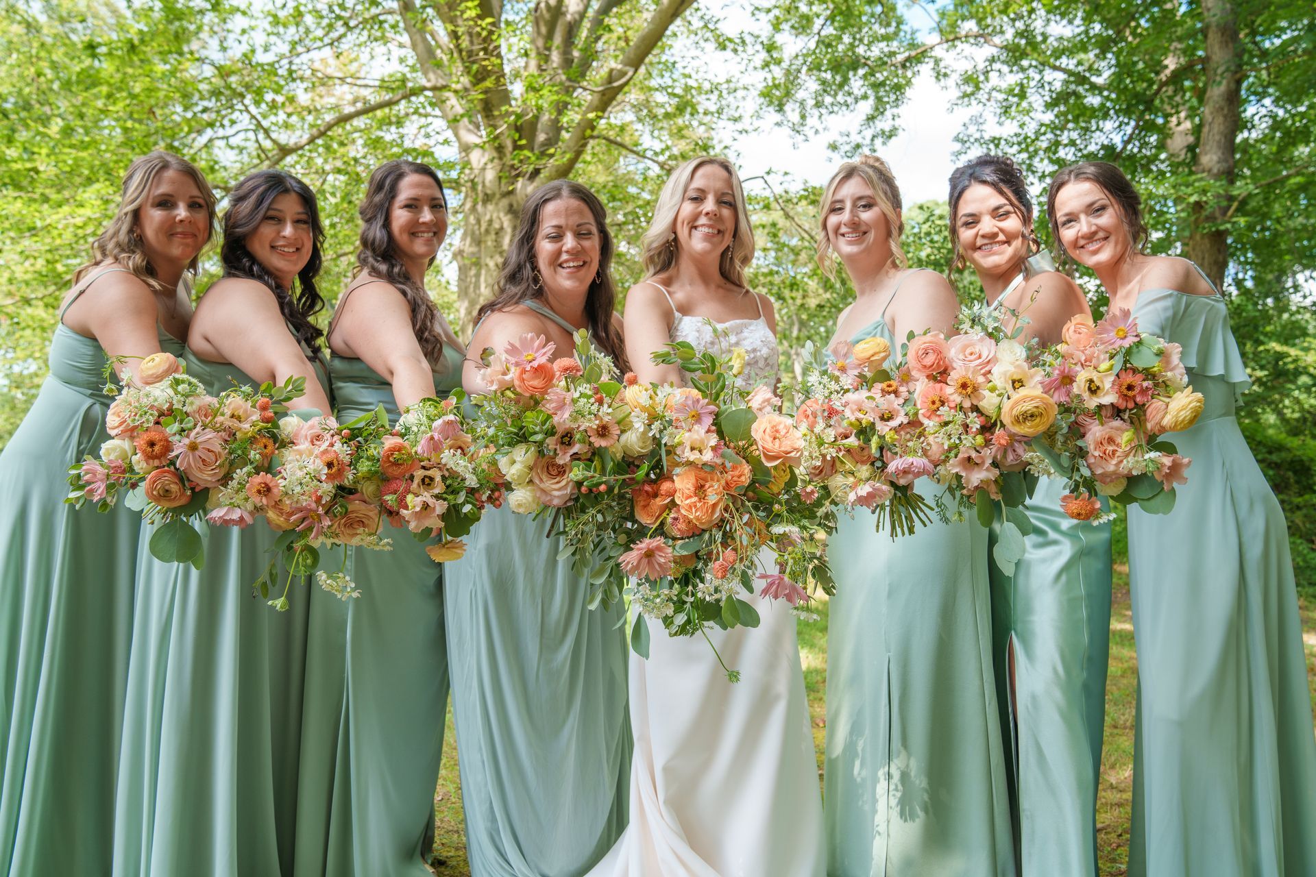 Bridesmaids in sage green dresses holding pastel bouquets outdoors in a garden setting