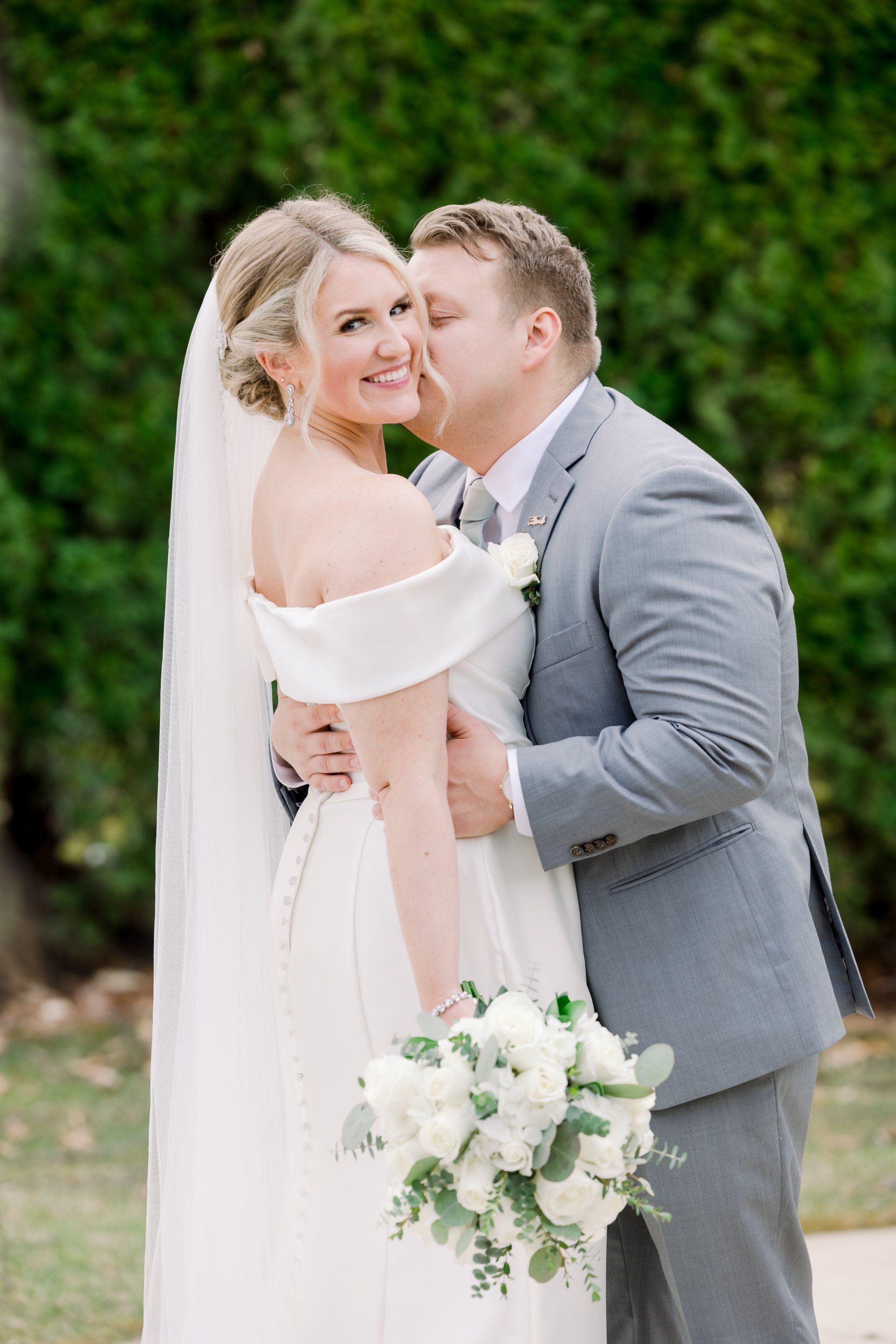 Bride and groom embracing outdoors, with the bride holding a white bouquet.