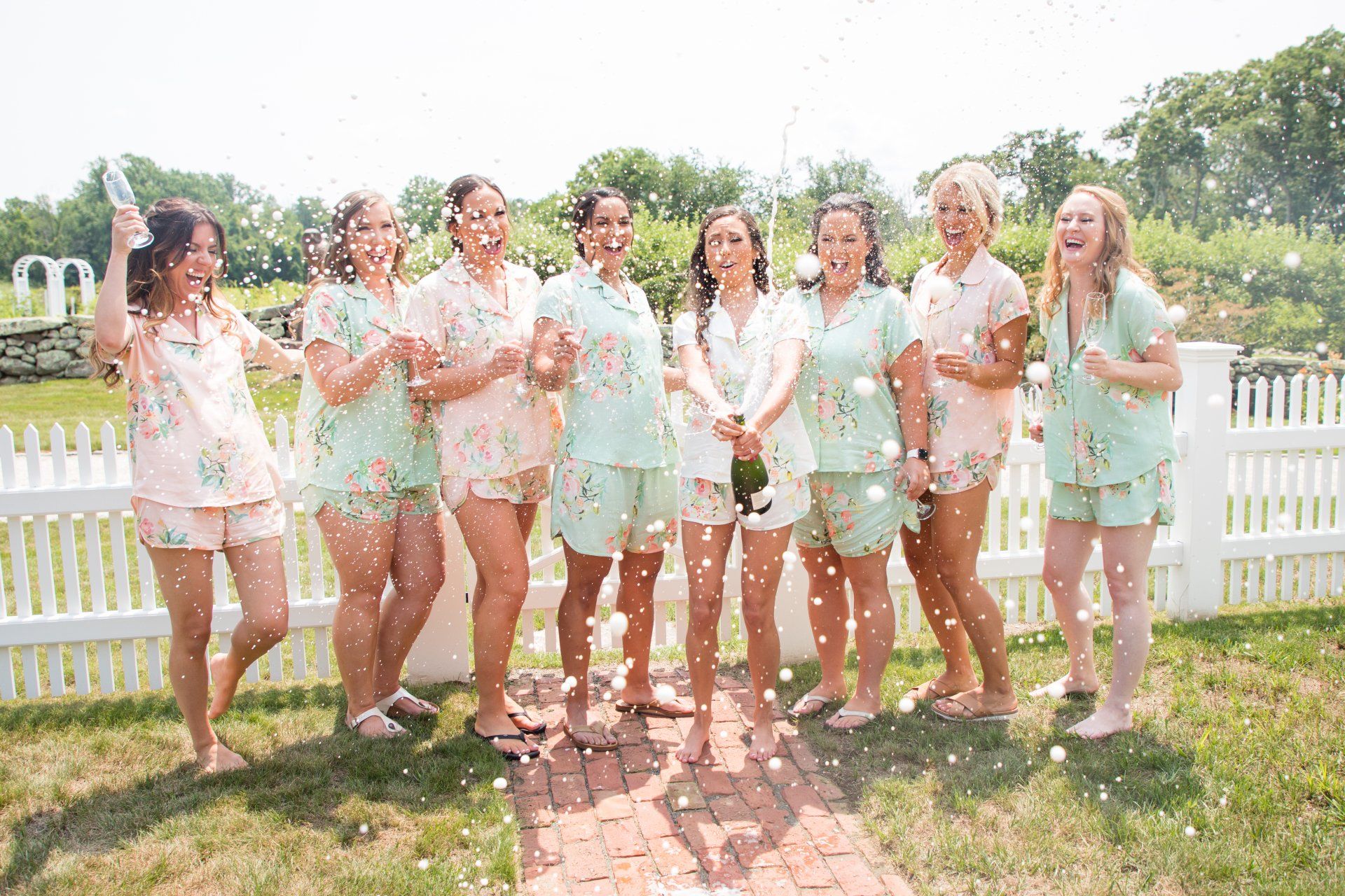 Group of people posing outdoors in light outfits, laughing in a sunny garden with a white fence and flowers.