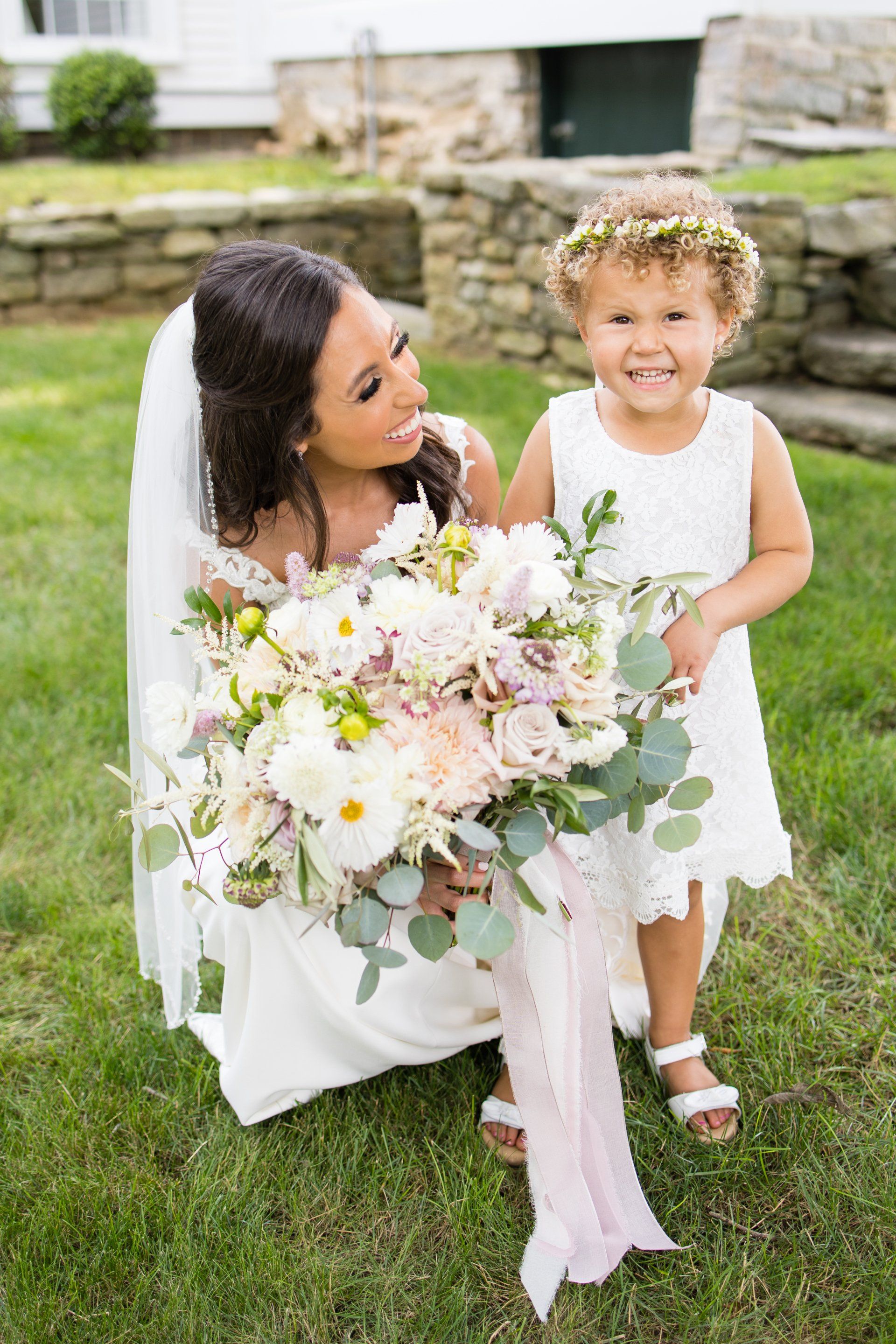 Woman in white kneels beside smiling child in white dress holding a large bouquet outdoors on grass