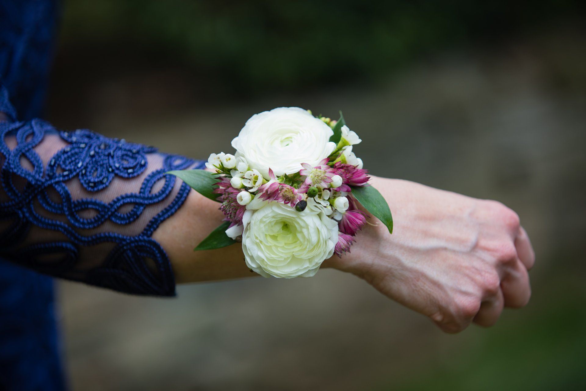 Wrist corsage of white and pink flowers with greenery on a person’s arm in a blue lace sleeve