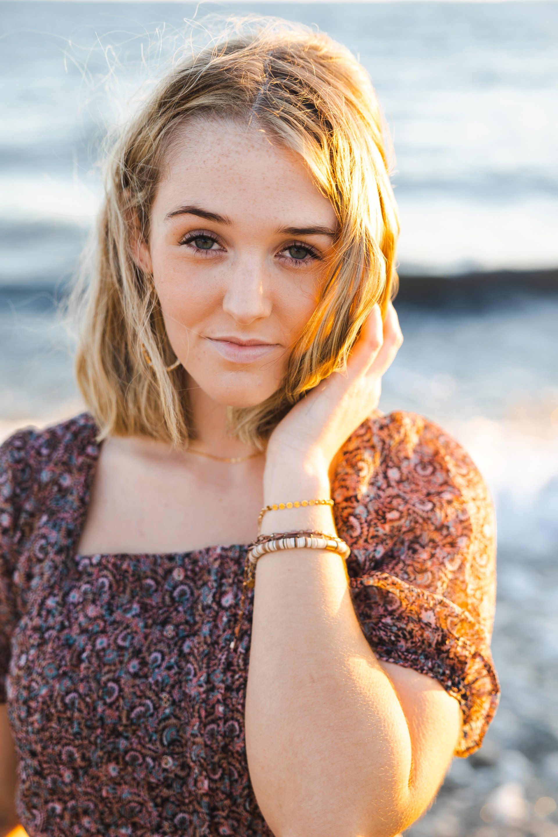 Woman standing by the water with one hand on her face, wearing a patterned top in warm sunset light